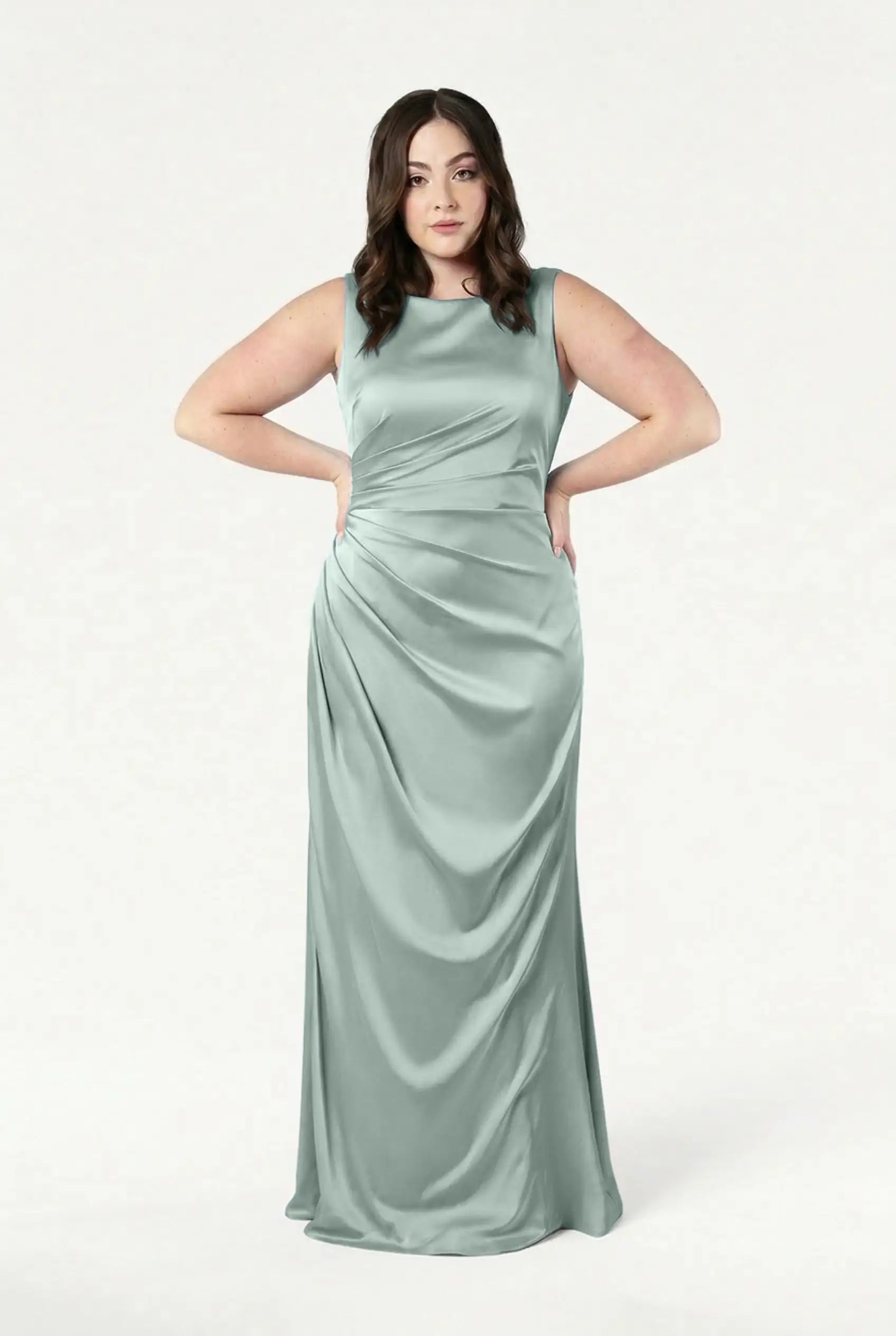 A woman poses confidently with her hands on her hips in the TH&TH Nala Bridesmaid Dress in Dusty Eucalyptus Satin, a size-inclusive, sleeveless, floor-length gown featuring gathered draping and Grecian-style pleating.