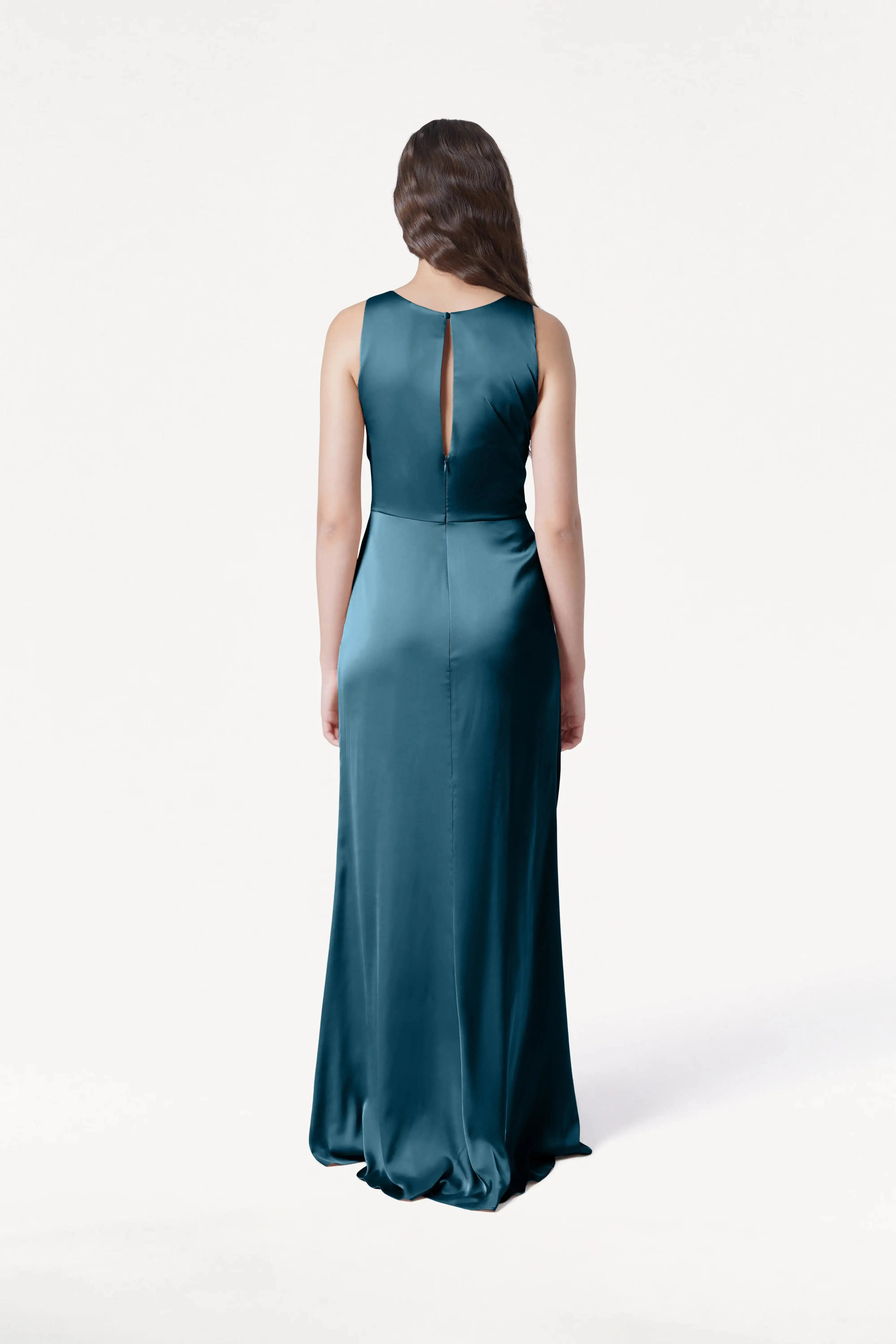A woman with long brown hair, wearing the TH&TH Nala Bridesmaid Dress in Emerald Dawn Satin, stands facing away against a plain white background.