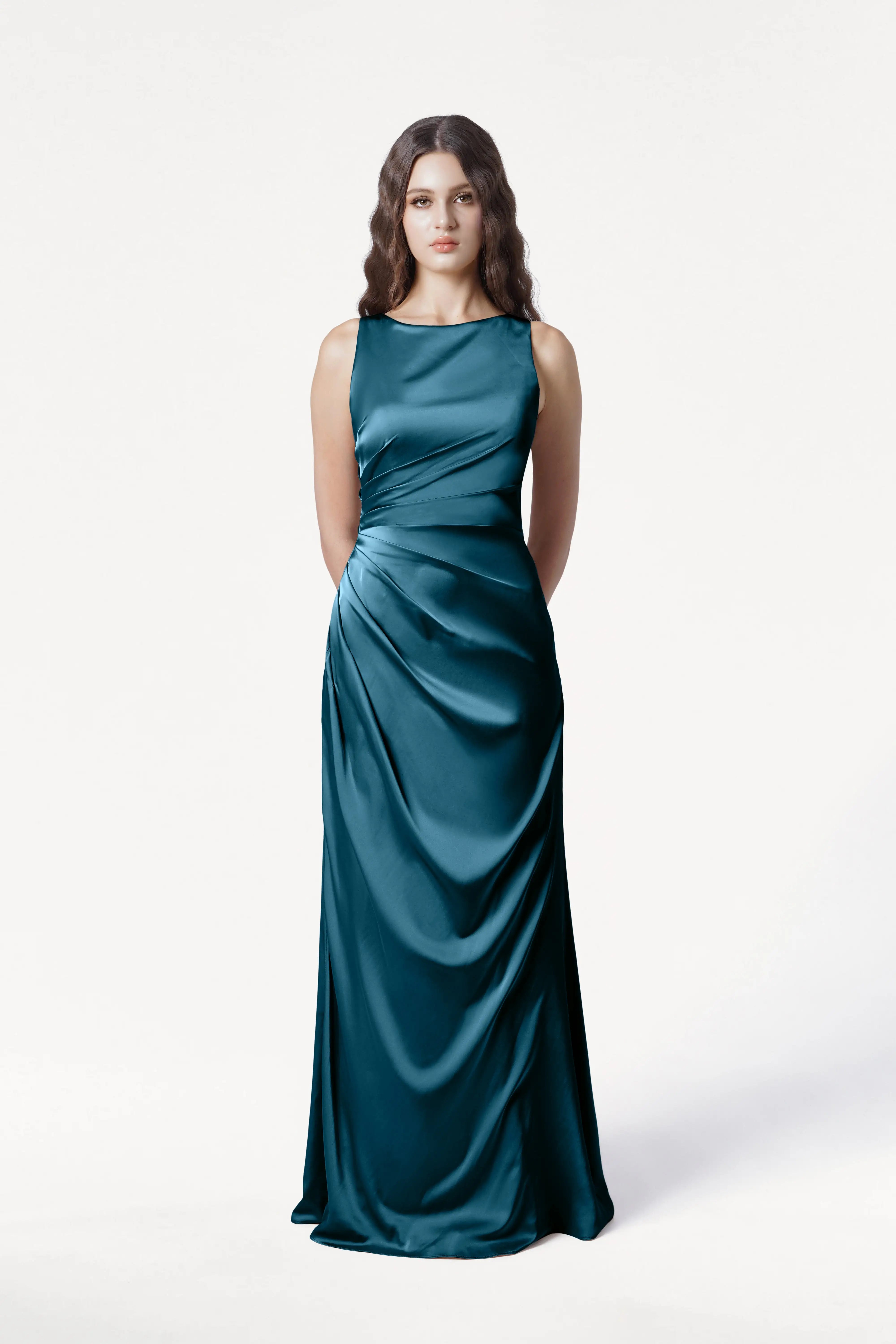 A woman with long wavy brown hair models the Nala Bridesmaid Dress in Emerald Dawn Satin by TH&TH—sleeveless and floor-length—standing against a plain white background, hands behind her back. Ideal for modern bridal parties.