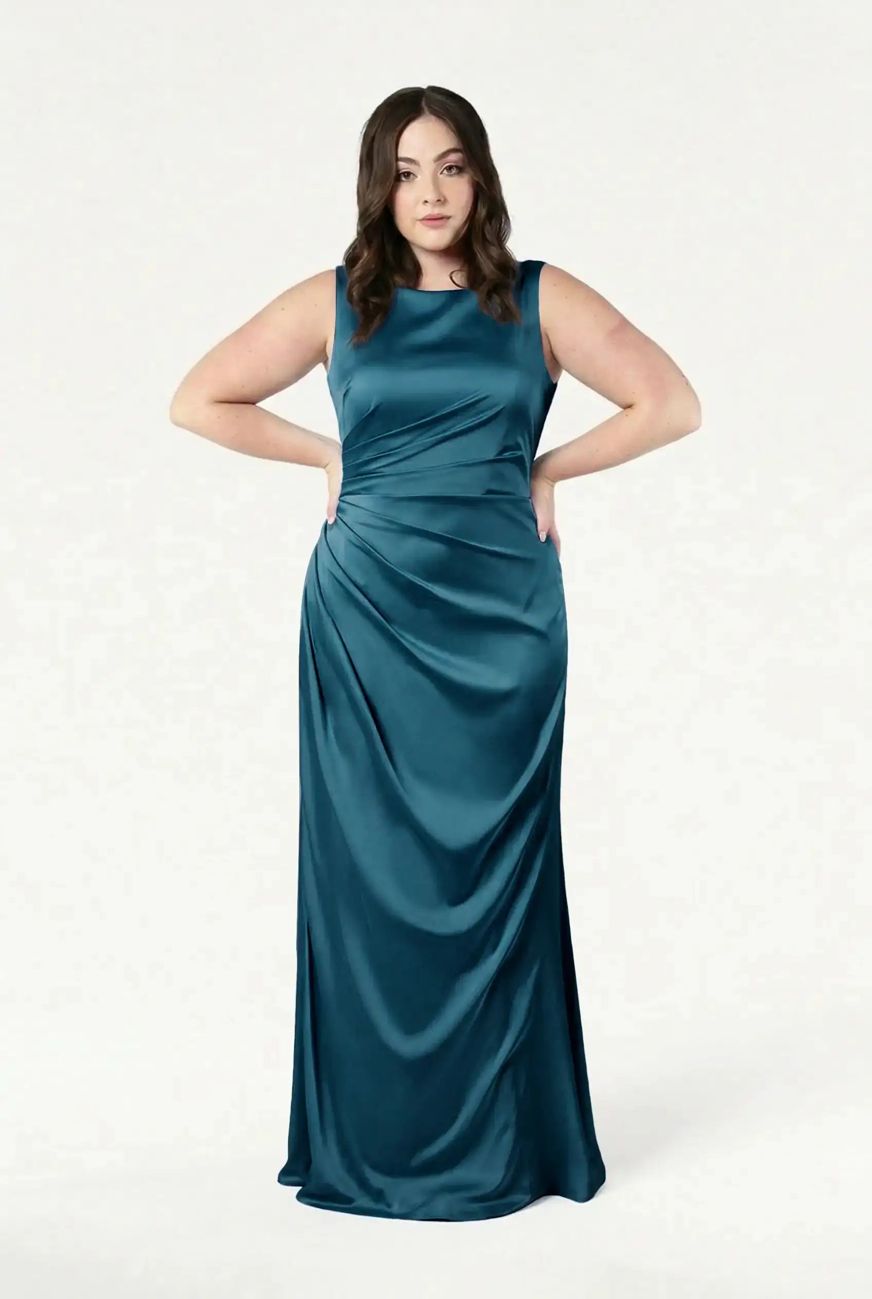 A woman with long brown hair stands hands on hips, modeling the TH&TH Nala Bridesmaid Dress in Emerald Dawn Satin, a sleeveless, floor-length gown with side ruching—ideal for modern bridal parties—against a plain white background.