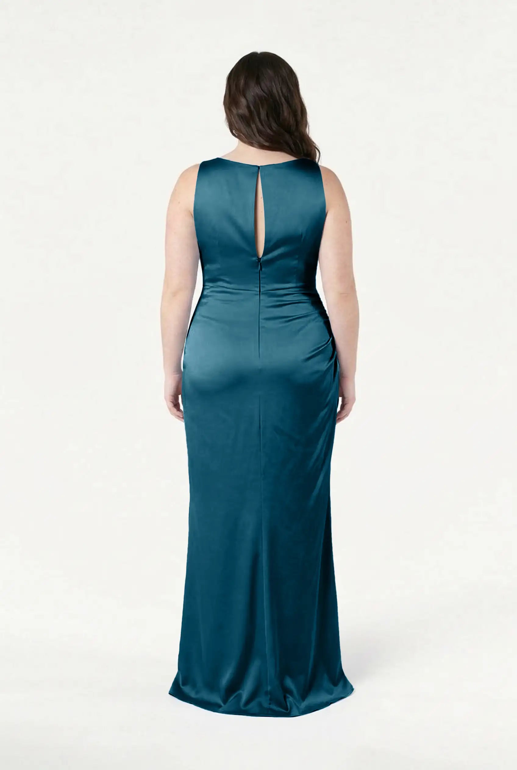 A woman faces away in the Nala Bridesmaid Dress in Emerald Dawn Satin by TH&TH—a floor-length, sleeveless gown with a keyhole back and fitted silhouette—ideal for modern bridal parties. The background is plain white.