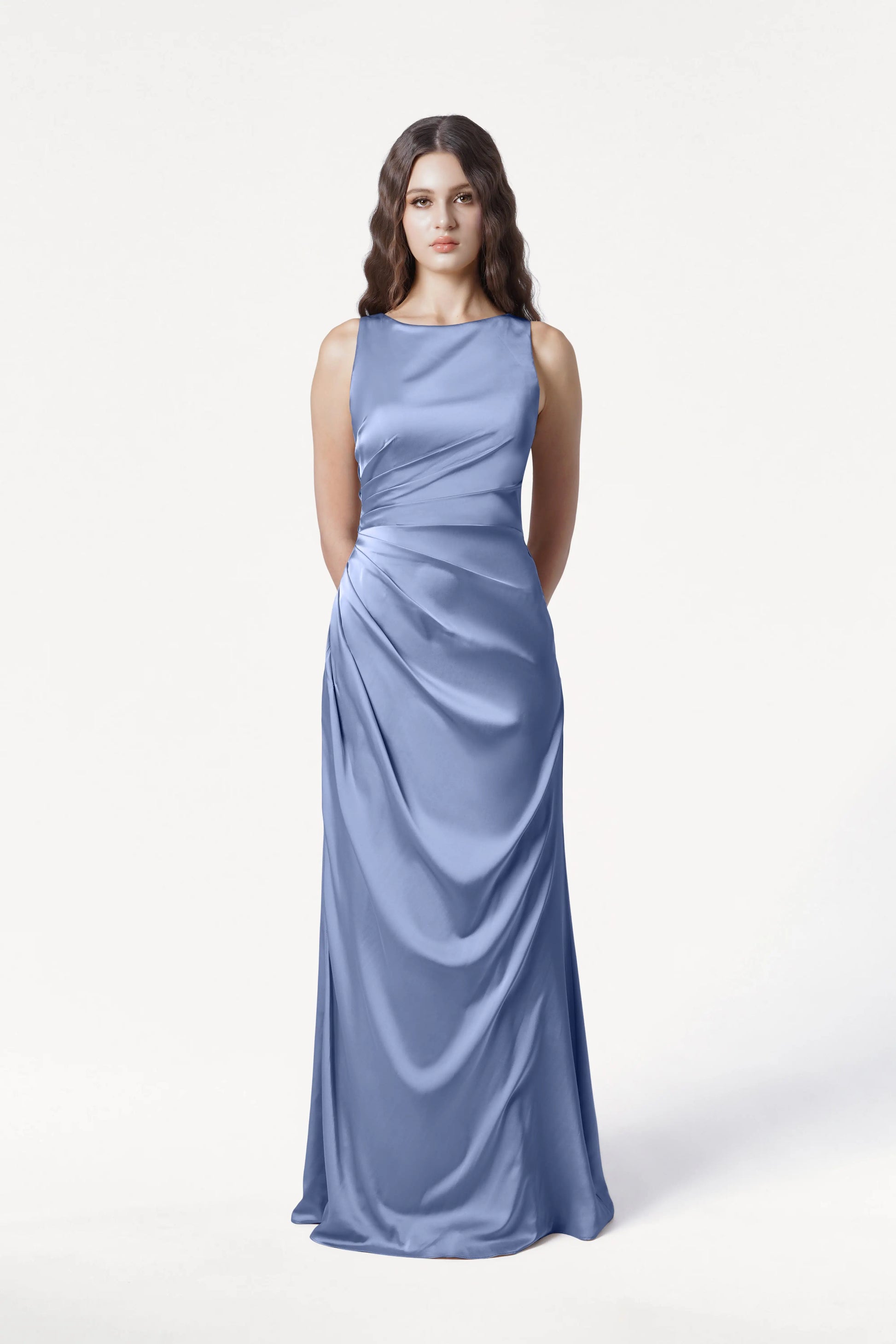 A woman with long brown hair in loose waves wears the TH&TH Nala Bridesmaid Dress in French Blue Satin, a size-inclusive, floor-length gown featuring Grecian pleating at the waist, posed against a plain white background.