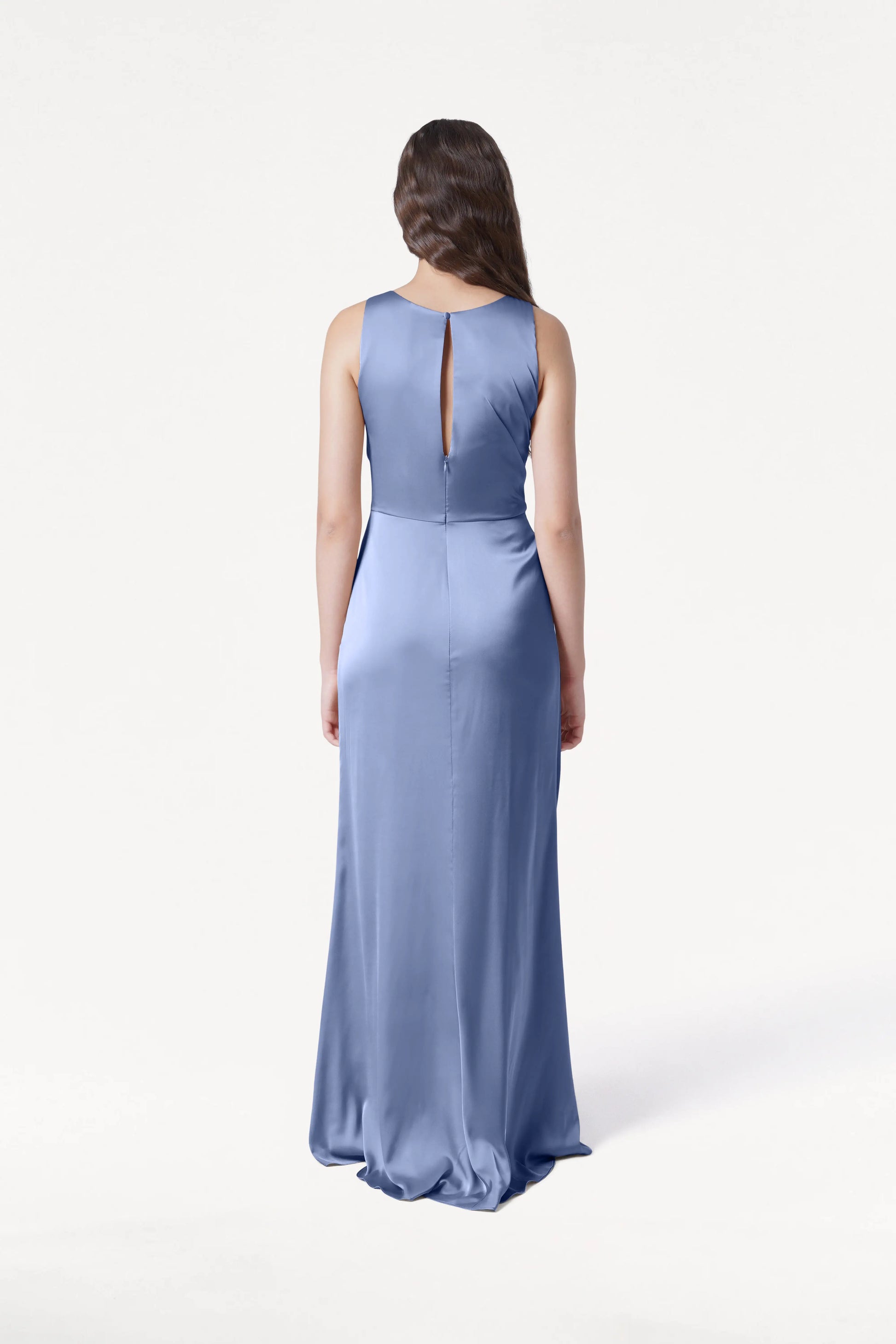 A woman with long brown hair, wearing the TH&TH Nala Bridesmaid Dress in French Blue Satin, featuring Grecian-inspired pleating and a sleeveless, floor-length design, stands facing away against a plain white background.