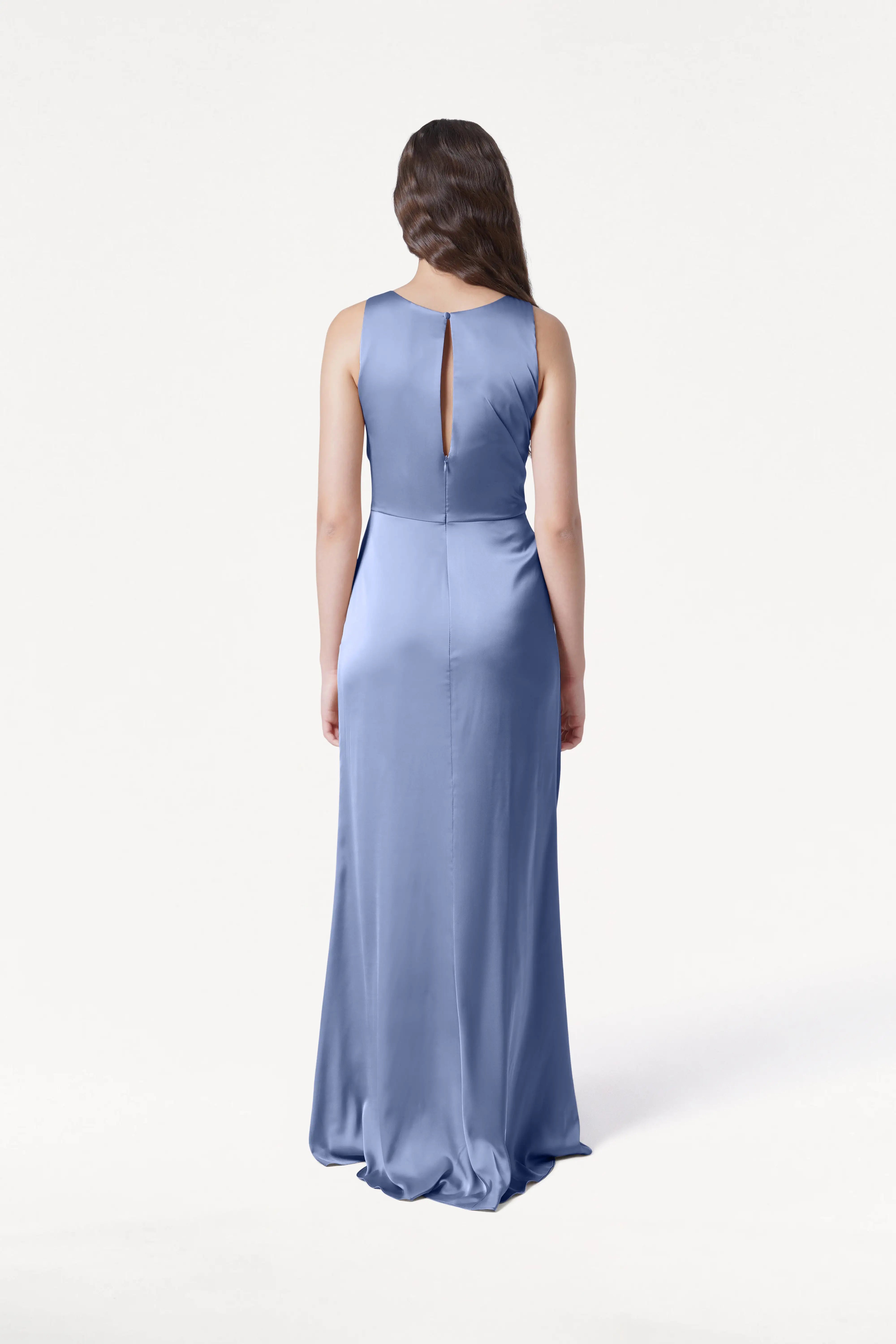 A woman with long brown hair, wearing the TH&TH Nala Bridesmaid Dress in French Blue Satin, featuring Grecian-inspired pleating and a sleeveless, floor-length design, stands facing away against a plain white background.