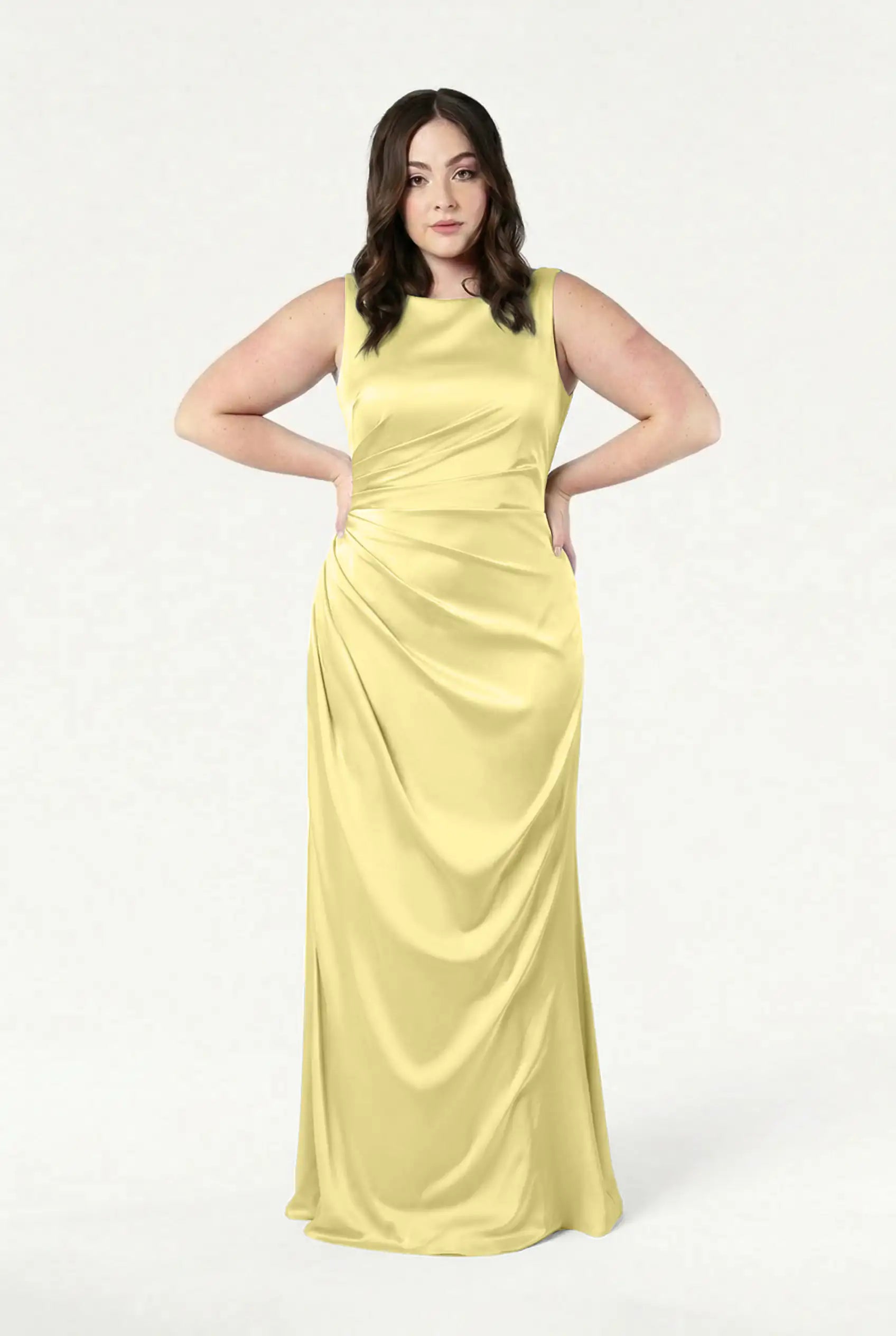 A woman with wavy brown hair stands hands-on-hips against a white background, wearing the size-inclusive TH&TH Nala Bridesmaid Dress in Limoncello Satin—a sleeveless, floor-length yellow gown.