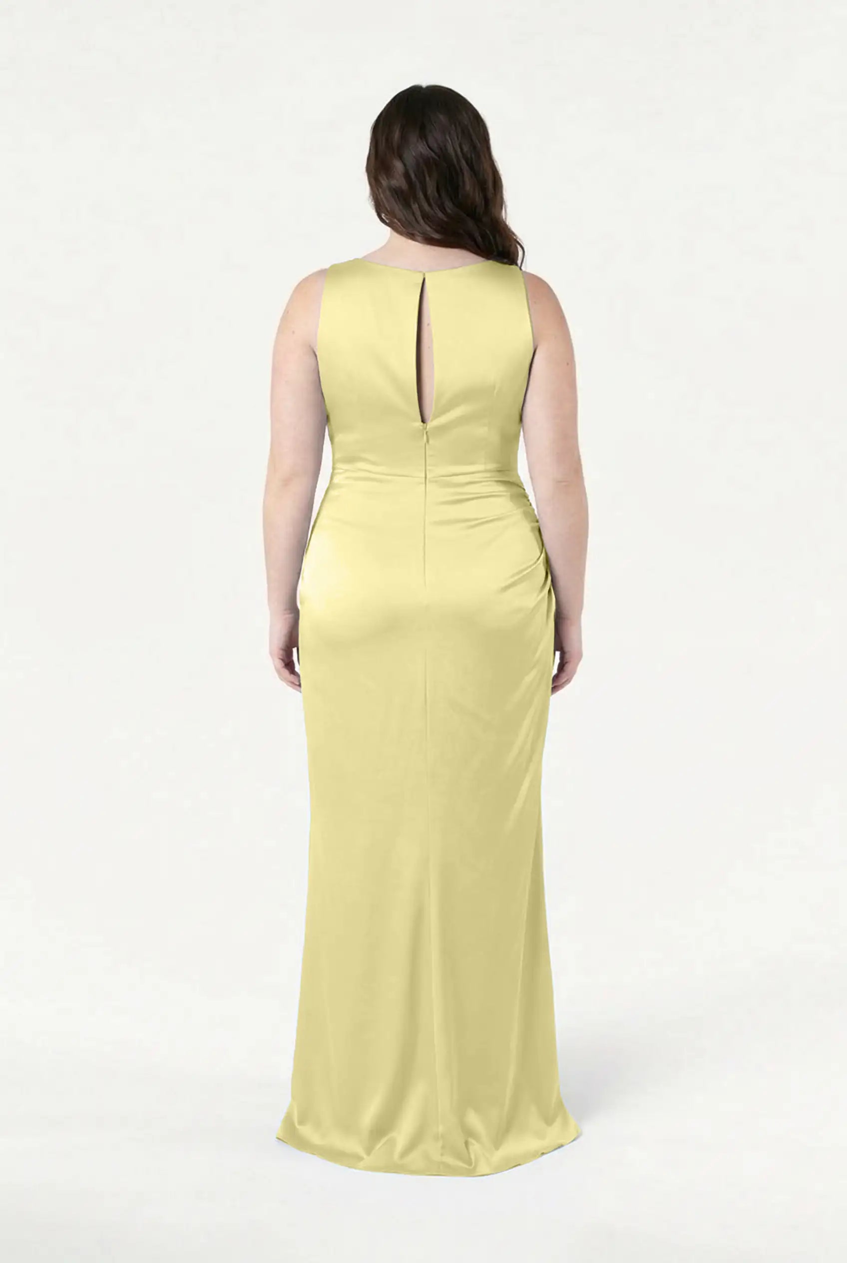 A woman with wavy brown hair is shown from behind wearing the TH&TH Nala Bridesmaid Dress in Limoncello Satin, a size-inclusive, floor-length gown with Grecian pleating and a keyhole back. The background is plain white.
