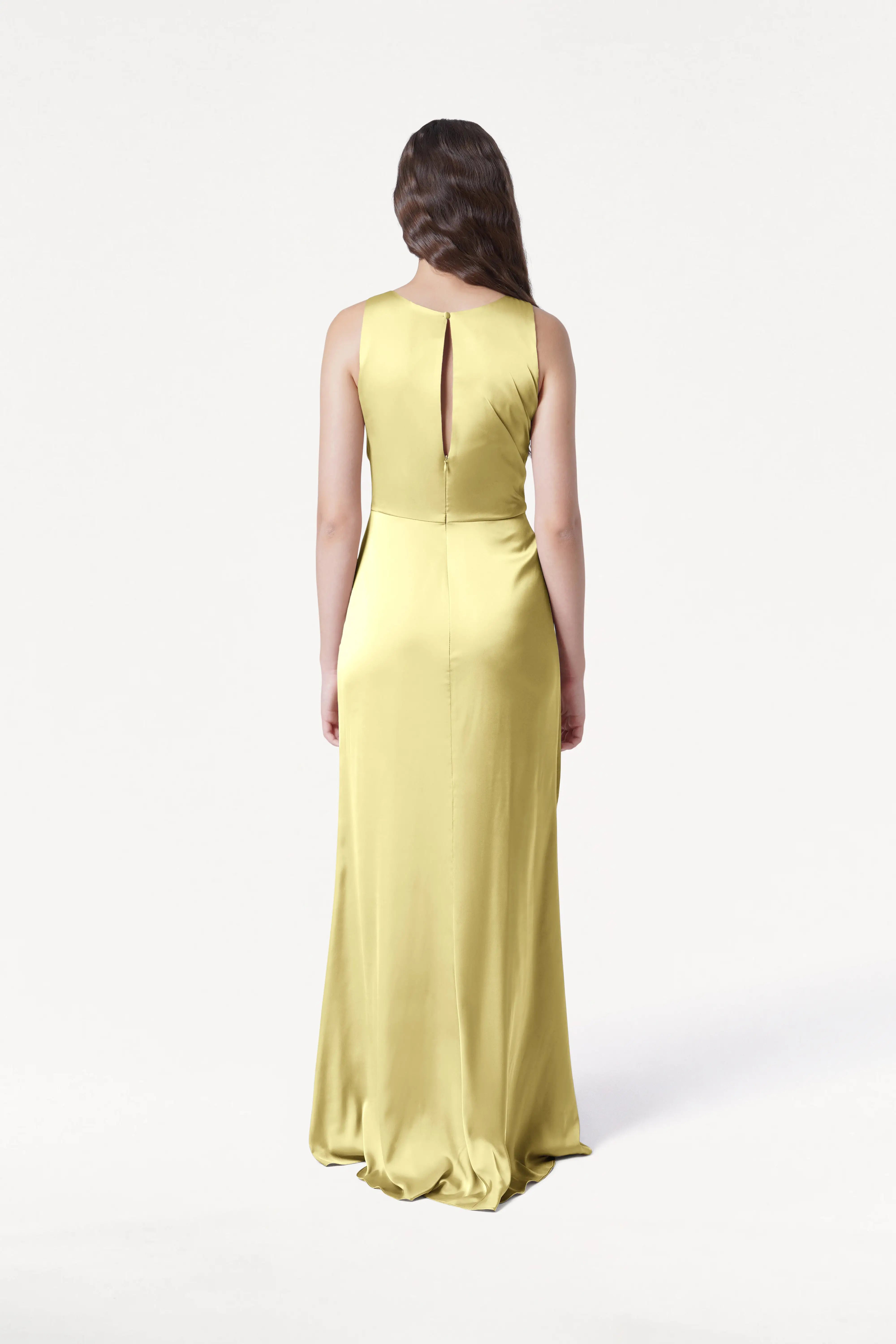 A woman with long brown hair stands facing away in the TH&TH Nala Bridesmaid Dress in Limoncello Satin, featuring size-inclusive fit, Grecian pleating, and a keyhole back. The background is plain white.