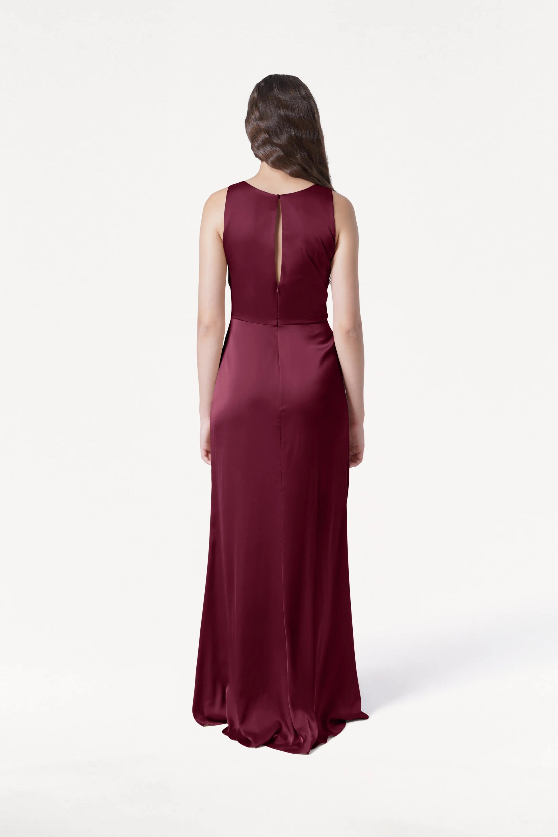 A woman with long brown hair faces away in the TH&TH Nala Bridesmaid Dress in Merlot Satin, a floor-length, size-inclusive gown with a sleeveless design and keyhole back, set against a plain white background.