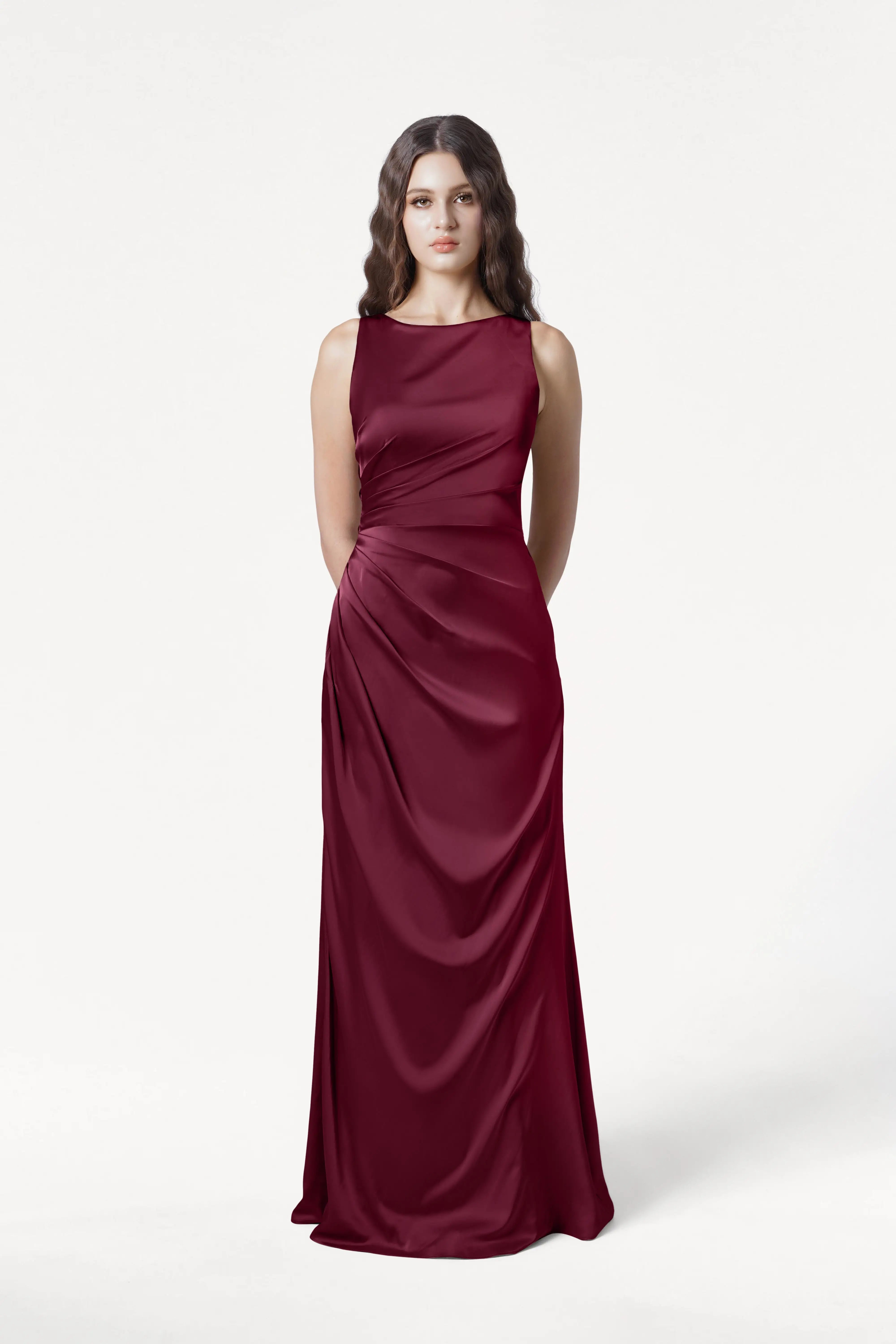 A woman with long wavy brown hair wears the TH&TH Nala Bridesmaid Dress in Merlot Satin, featuring Grecian-inspired pleating and a sleeveless, floor-length silhouette. She stands facing forward against a plain white background.