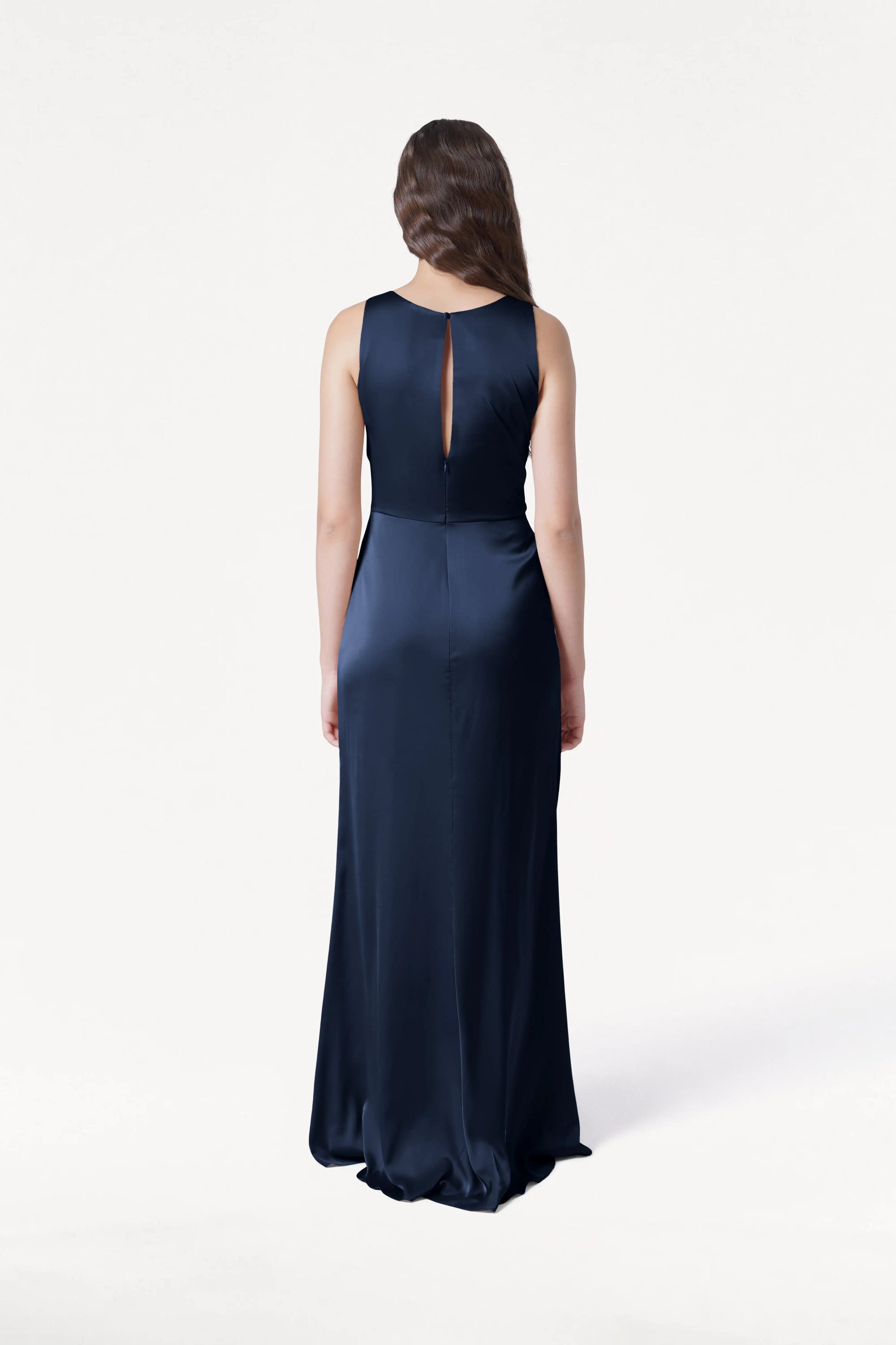 A woman with long brown hair stands facing away, wearing the TH&TH Nala Bridesmaid Dress in navy satin with a sleeveless, floor-length design and small back keyhole, set against a plain white background.