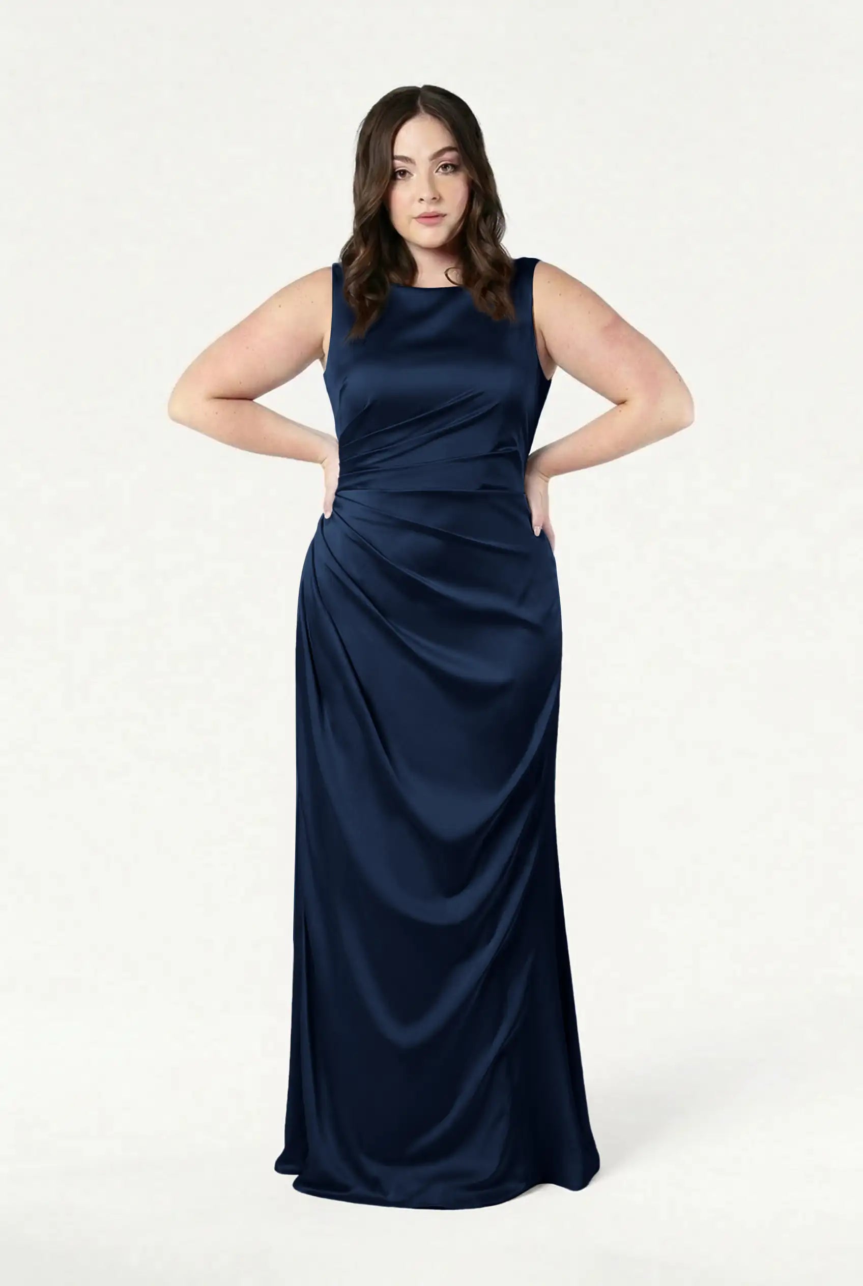 A woman with long brown hair stands against a white background, wearing the TH&TH Nala Bridesmaid Dress in navy satin with ruched detailing, her hands resting on her hips.