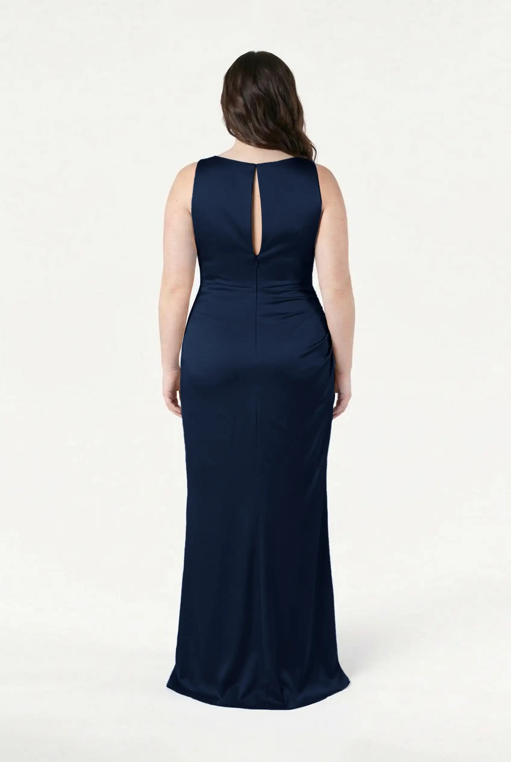A woman faces away in the TH&TH Nala Bridesmaid Dress in Navy Satin, showcasing its sleeveless, floor-length design and back keyhole detail against a plain white background.