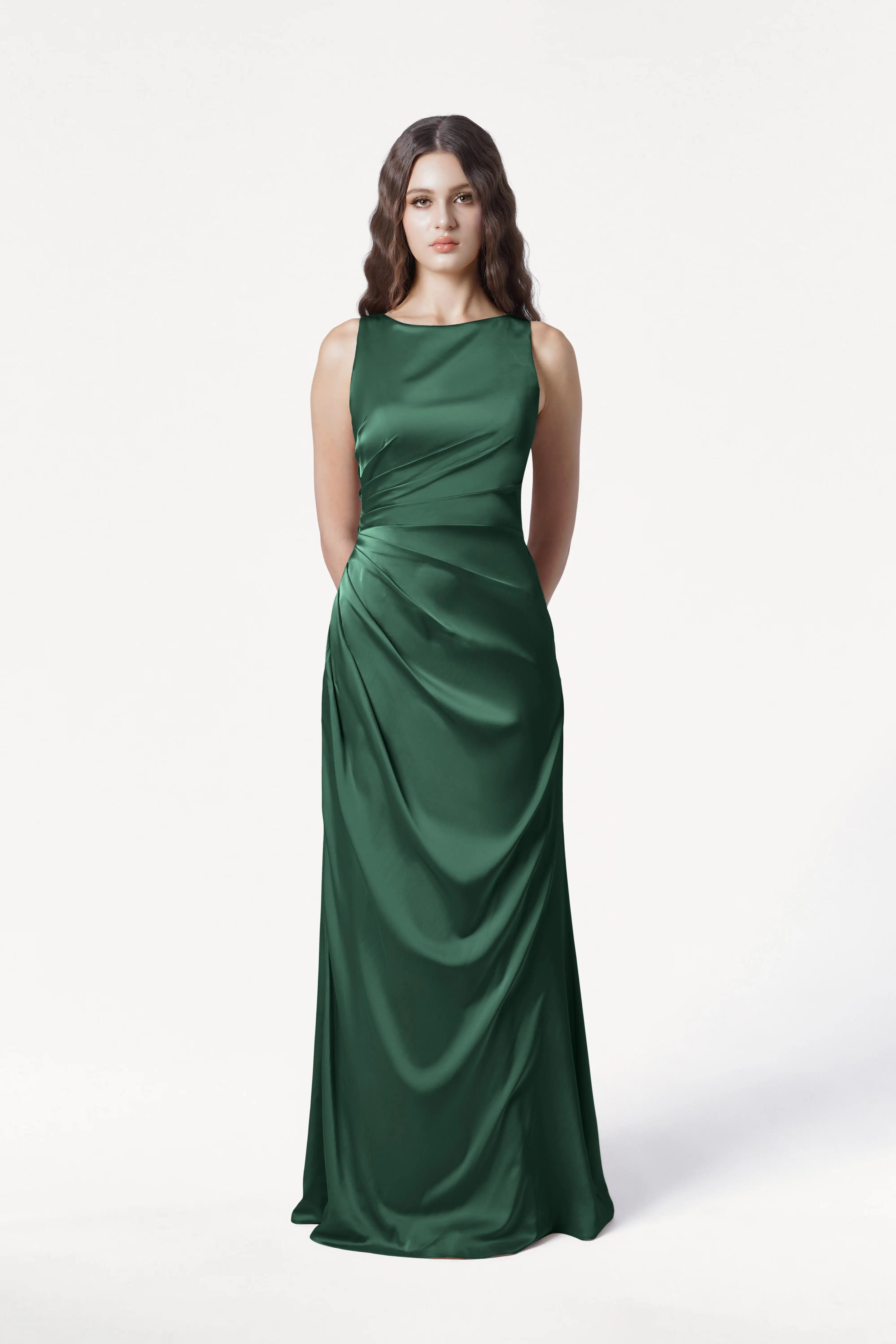 A woman with long wavy hair stands against a white background, wearing the sleeveless, floor-length Nala Bridesmaid Dress in Olive Grove Satin by TH&TH, featuring gathered detail on one side—an elegant choice for modern bridal parties. Her hands are behind her back.