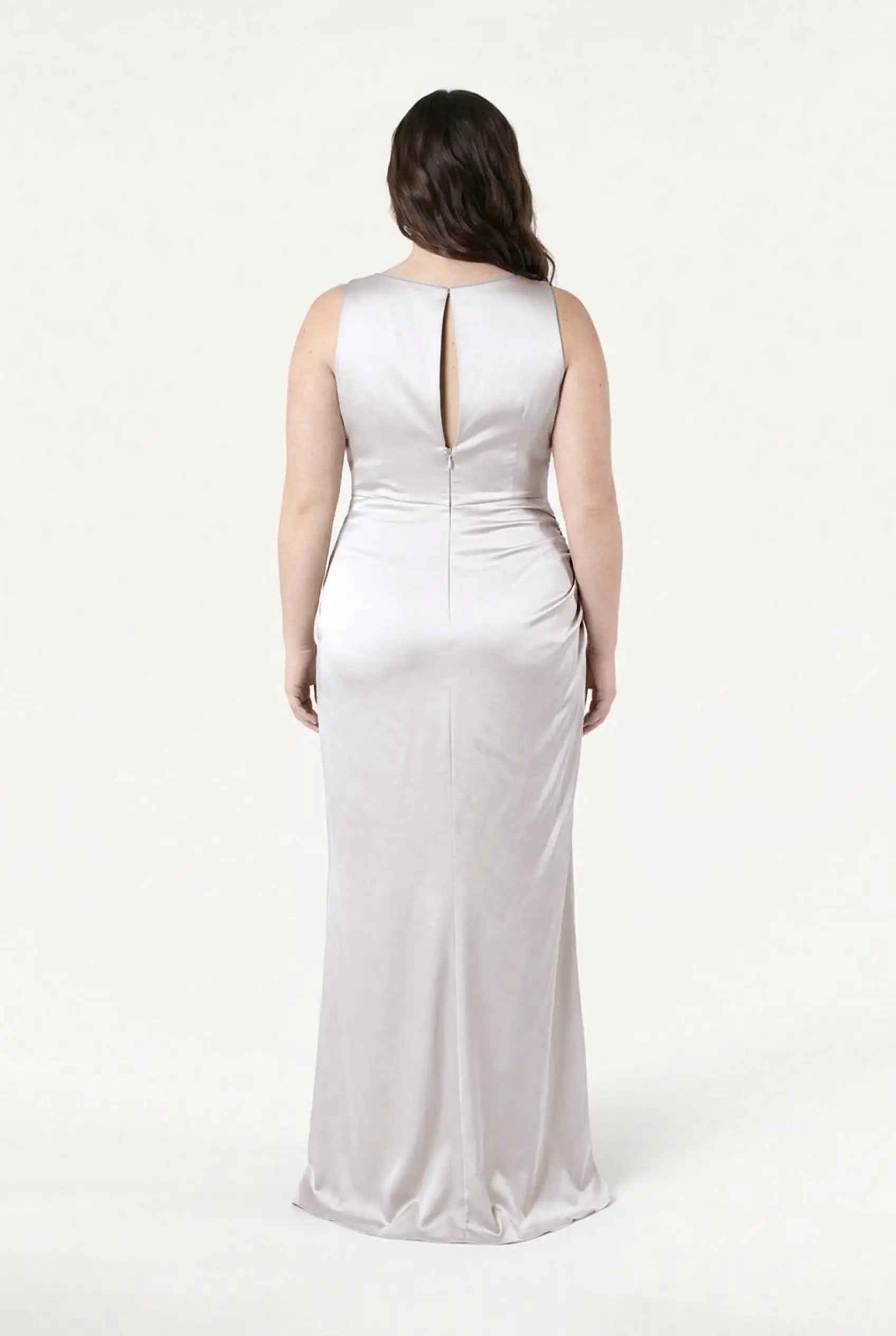 A woman with long dark hair wears the TH&TH Nala Bridesmaid Dress in Oyster Satin, featuring a sleeveless, floor-length silhouette with Grecian pleats and a back keyhole cutout elegantly displayed to the camera.