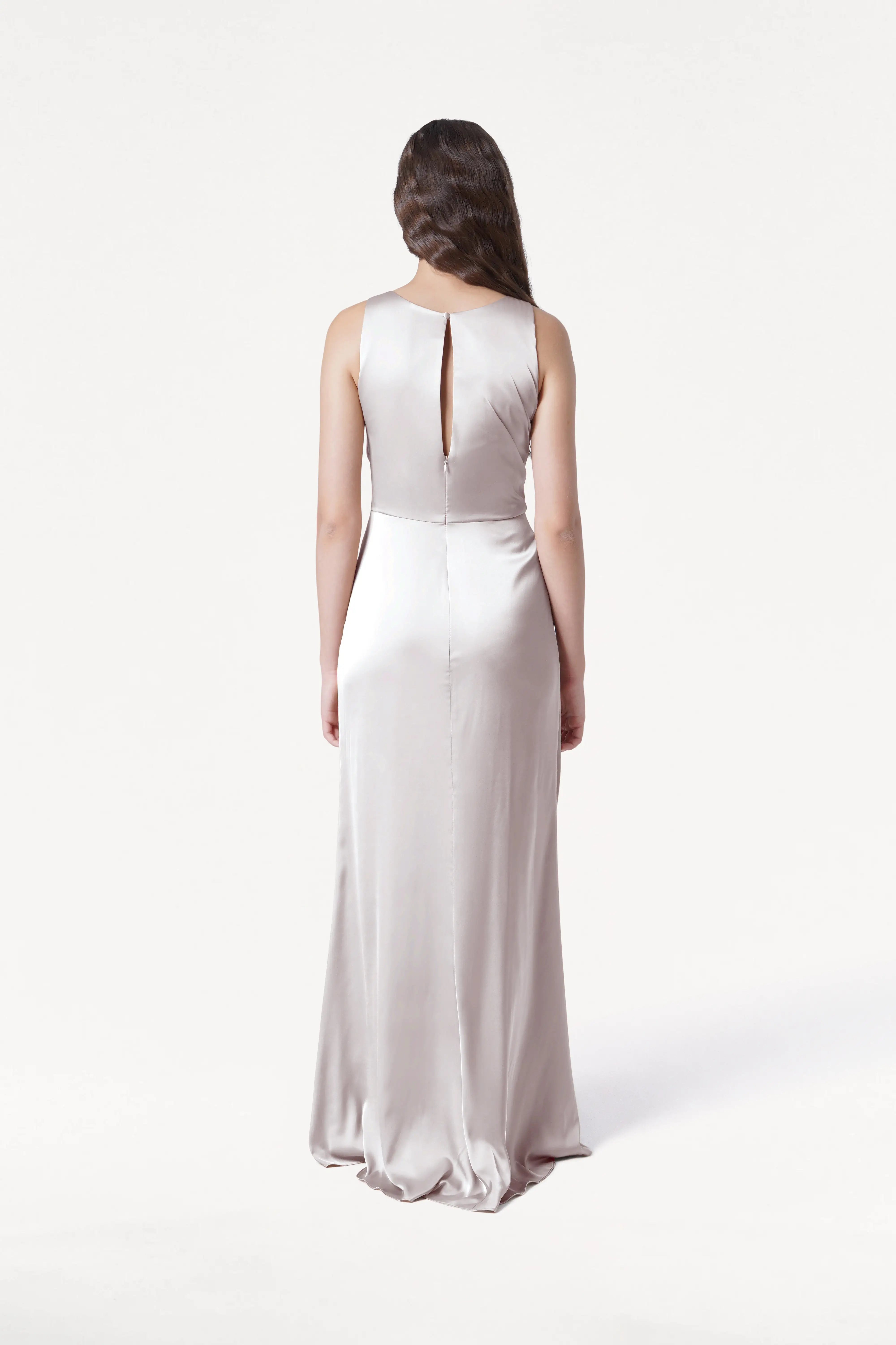 A woman with long brown hair faces away, wearing the TH&TH Nala Bridesmaid Dress in Oyster Satin—a floor-length, sleeveless, size-inclusive gown featuring Grecian pleats and a keyhole back—against a plain white background.