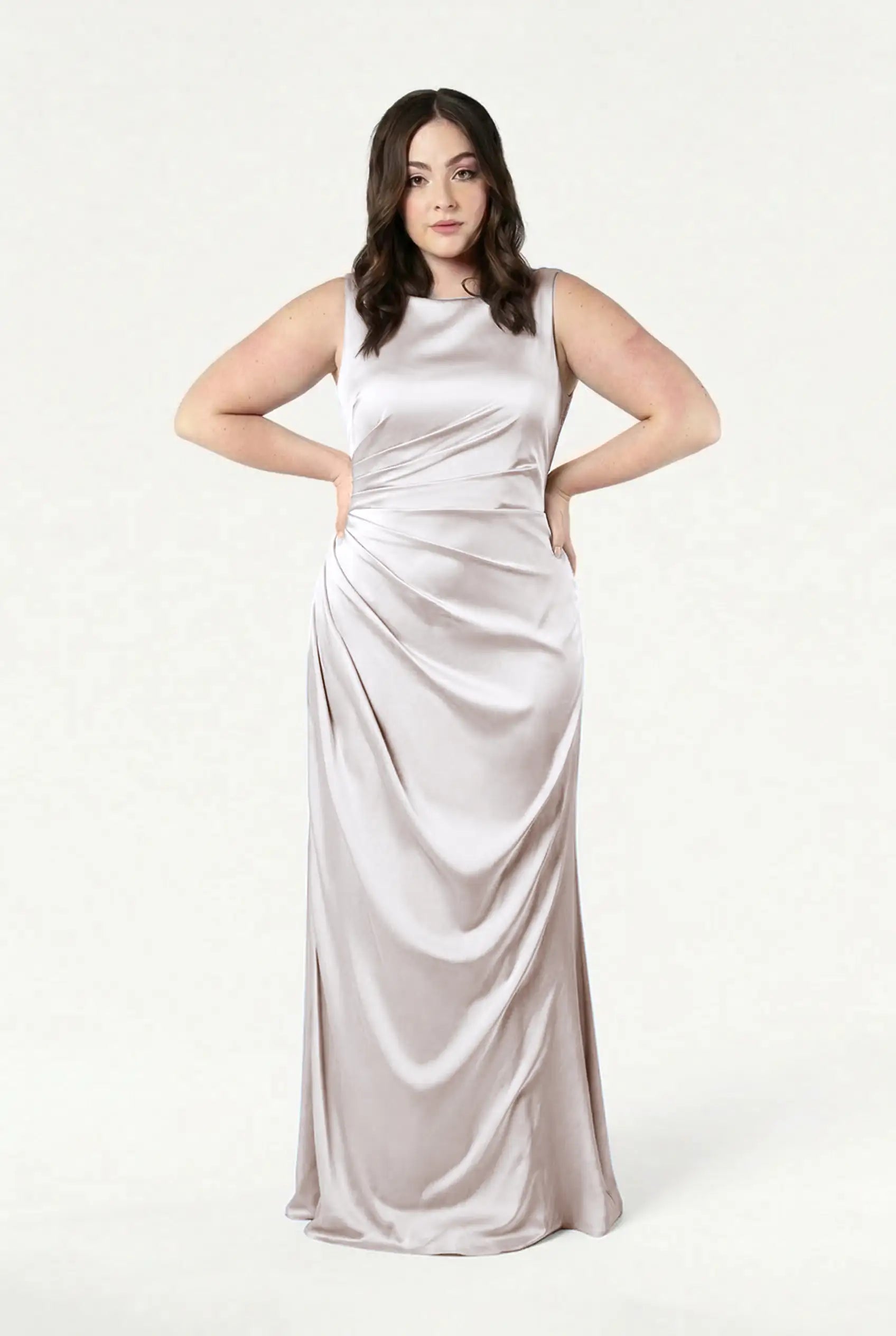 A woman with wavy brown hair models the TH&TH Nala Bridesmaid Dress in Oyster Satin, a sleeveless, floor-length gown with Grecian pleats and draped details, standing hands on hips against a plain white background.