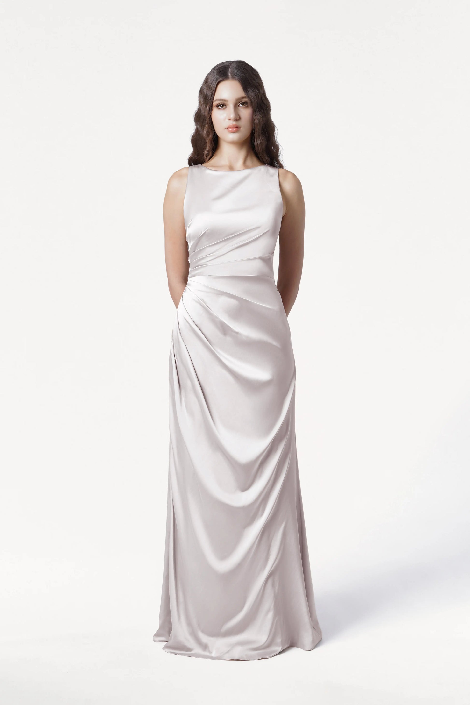 A woman with long wavy brown hair stands against a white background, wearing the TH&TH Nala Bridesmaid Dress in Oyster Satin— a sleeveless, floor-length gown with Grecian-inspired pleats and a fitted waist. Her hands are behind her back.