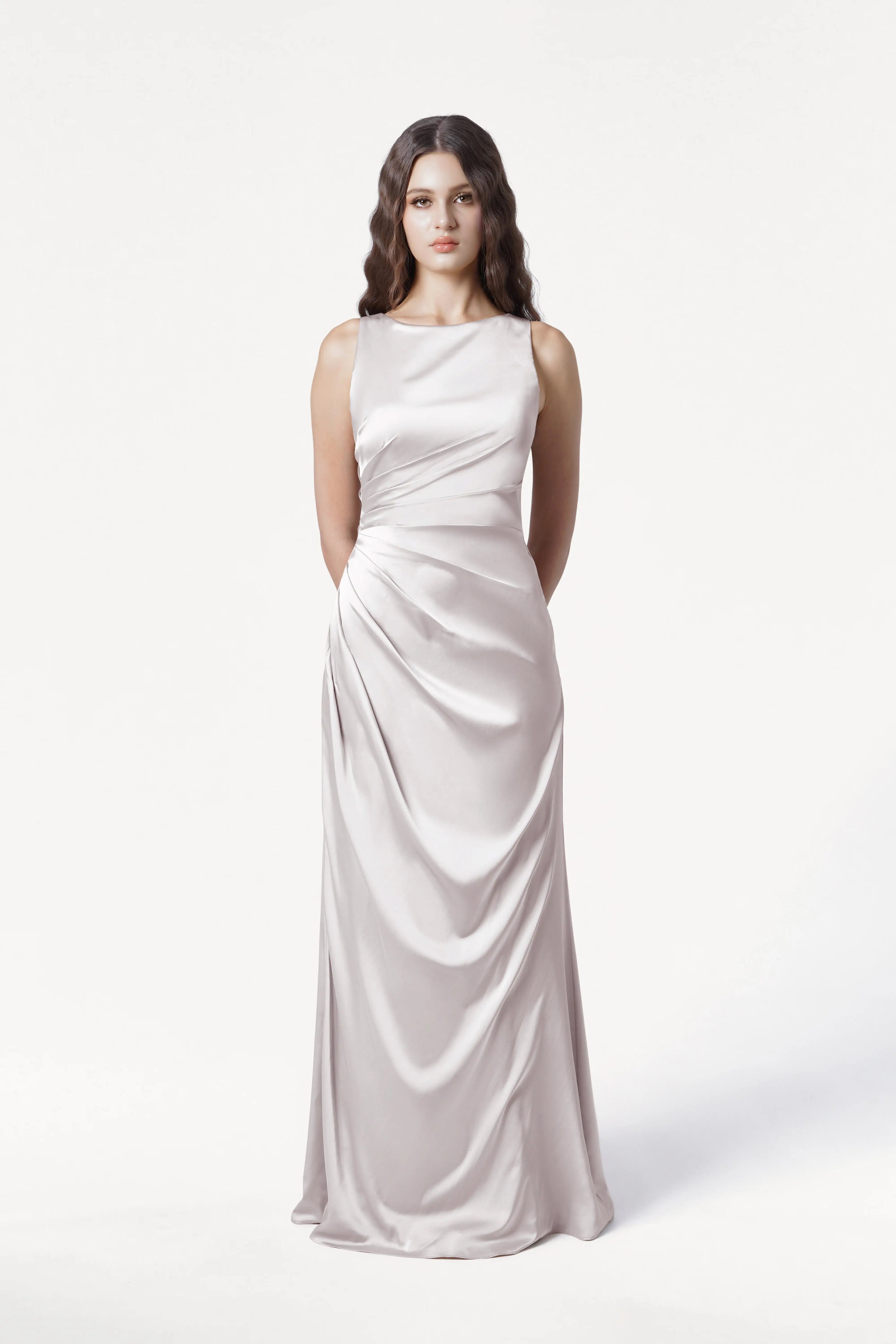 A woman with long wavy brown hair stands against a white background, wearing the TH&TH Nala Bridesmaid Dress in Oyster Satin— a sleeveless, floor-length gown with Grecian-inspired pleats and a fitted waist. Her hands are behind her back.