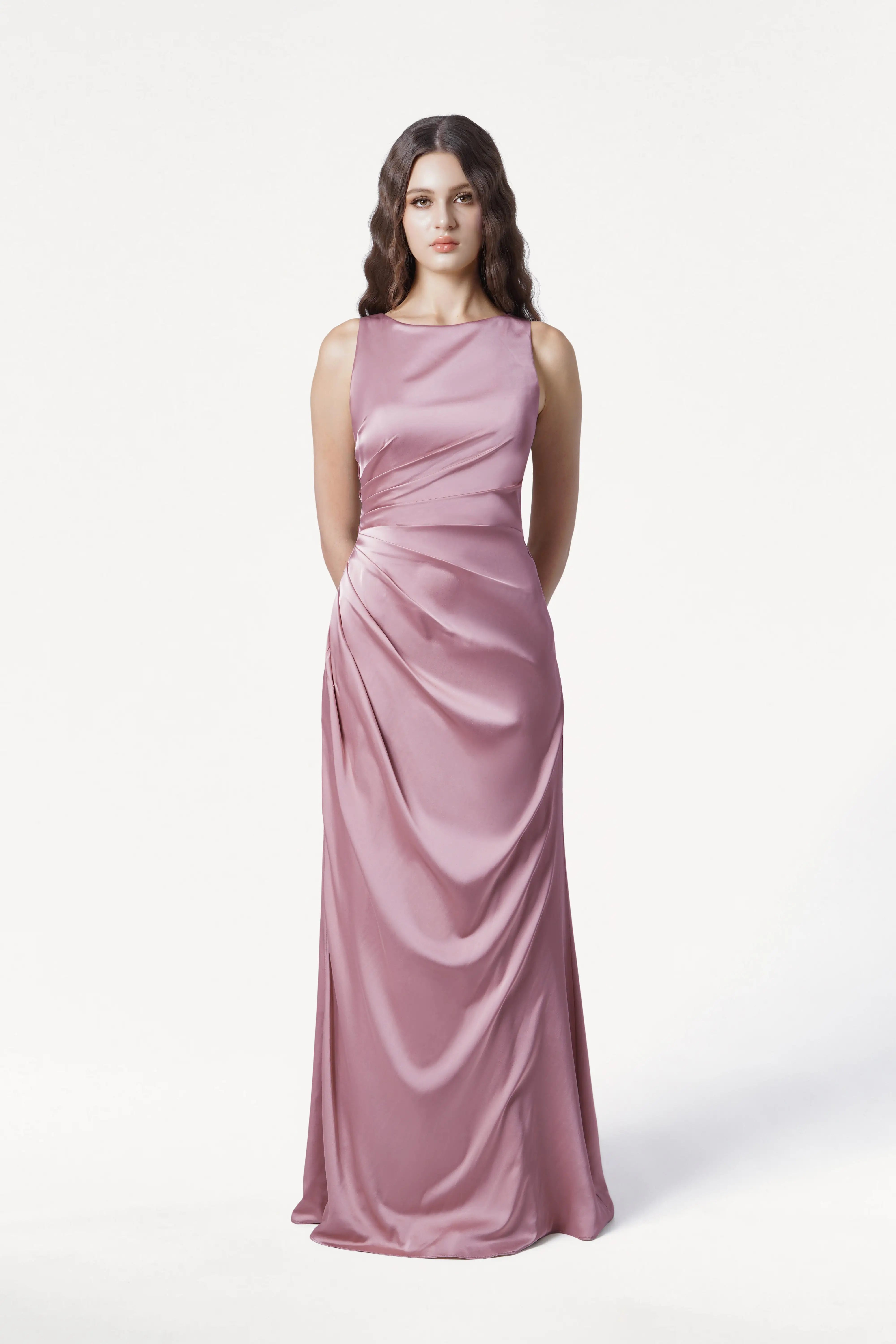 A woman faces forward against a white backdrop, modeling the floor-length, sleeveless Nala Bridesmaid Dress in Rose Pink Satin by TH&TH, featuring gentle side draping—an elegant choice for modern bridal parties. Her long wavy hair is worn down.