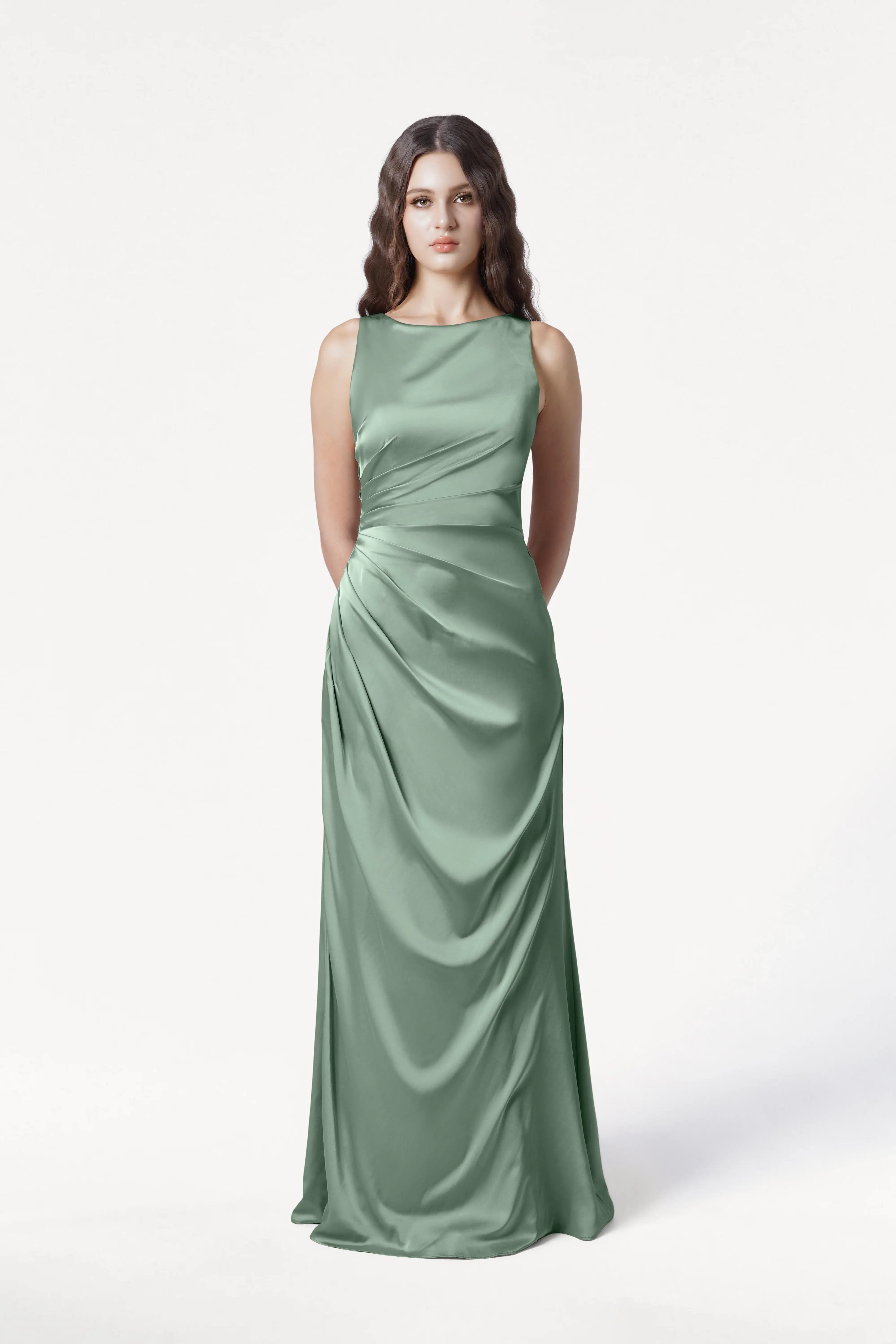 A woman with long wavy brown hair models the size-inclusive TH&TH Nala Bridesmaid Dress in sage green satin, a sleeveless floor-length gown with Grecian-inspired pleating, standing against a plain white backdrop.