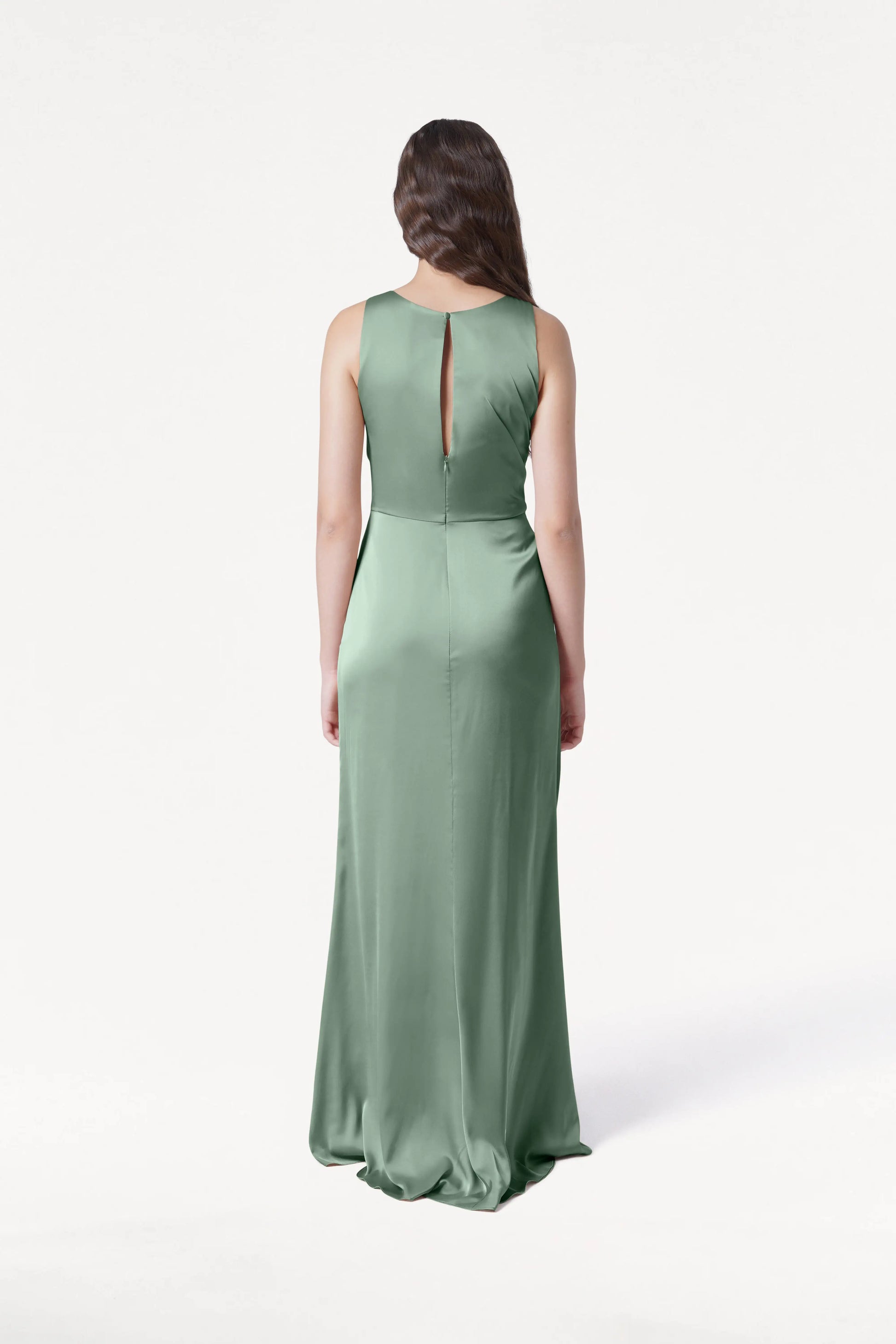 A woman is shown from the back wearing the TH&TH Nala Bridesmaid Dress in sage green satin, a size-inclusive, long, sleeveless dress with a keyhole upper back and softly flowing skirt against a plain white background.