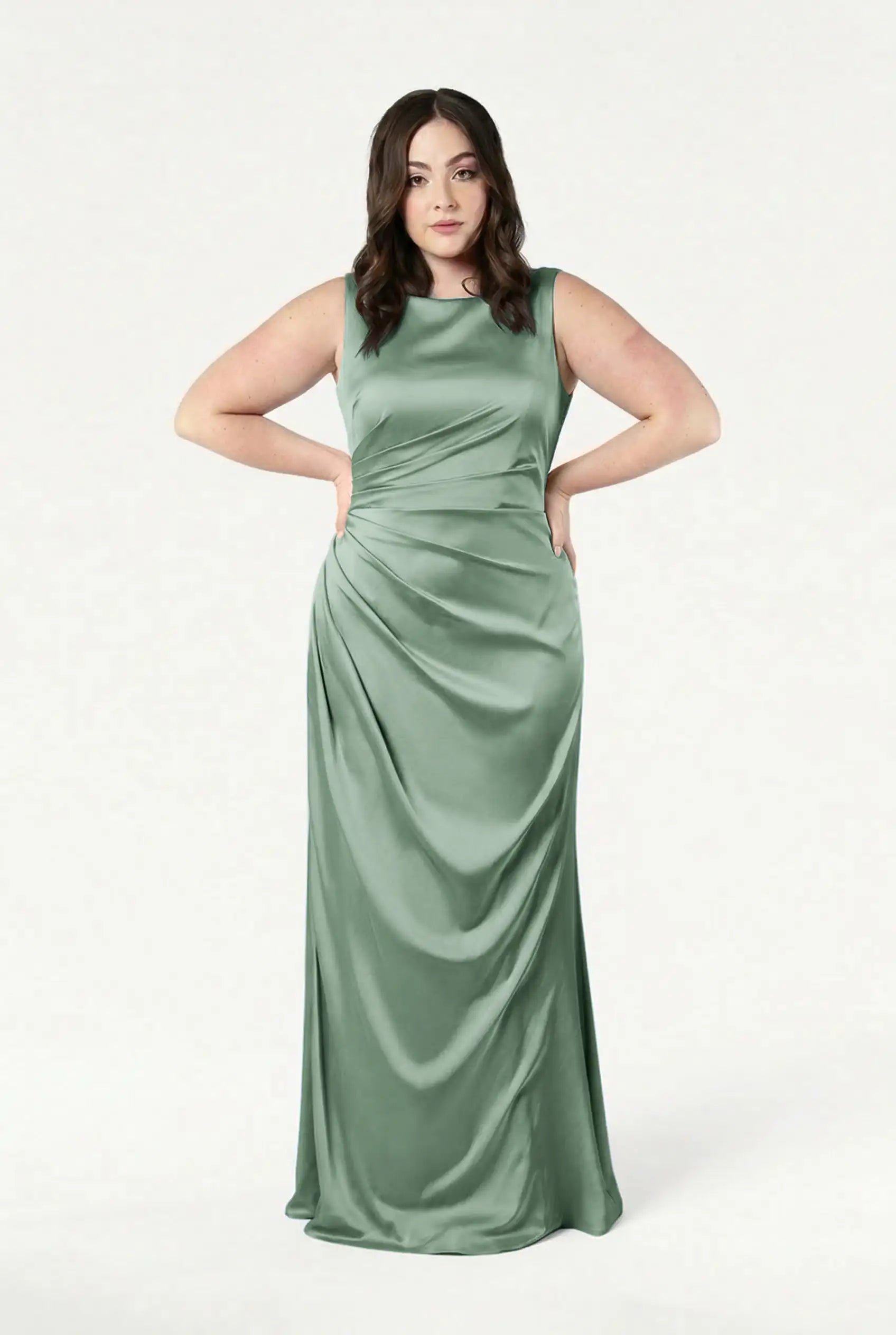 A woman stands with hands on hips, wearing the TH&TH Nala Bridesmaid Dress in sage green satin—a size-inclusive, sleeveless, floor-length gown with a softly draped skirt and Grecian-inspired pleating—against a plain white background.