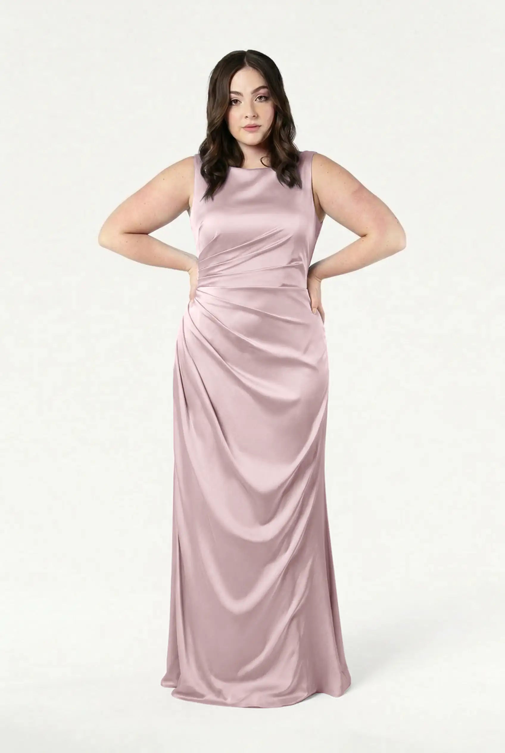 A woman with wavy brown hair poses against a white background in the TH&TH Nala Bridesmaid Dress in Smoked Blush Satin, a sleeveless, floor-length gown with Grecian-style pleating and ruching—ideal for modern weddings. Her hands rest on her hips.