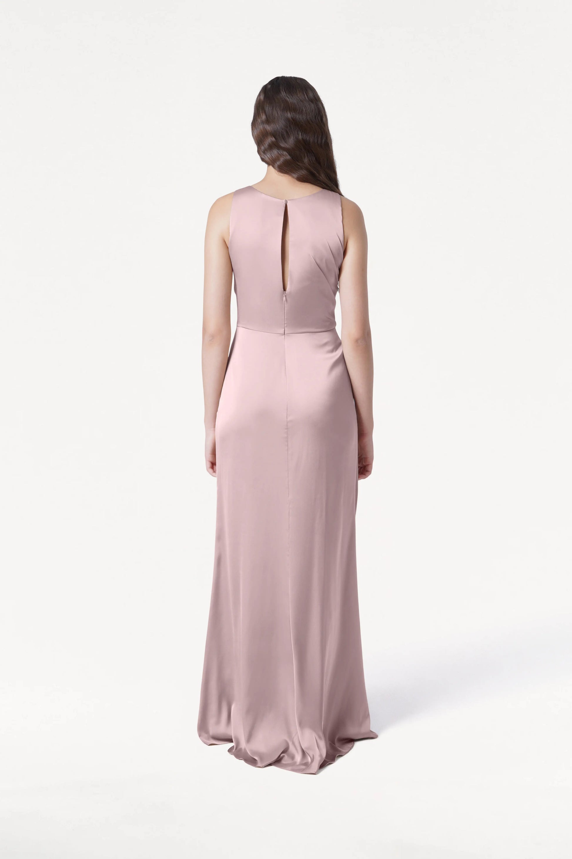 A woman with long brown hair faces away, wearing the TH&TH Nala Bridesmaid Dress in smoked blush satin. This sleeveless, floor-length gown features Grecian-inspired pleating and a keyhole back. The background is plain white.