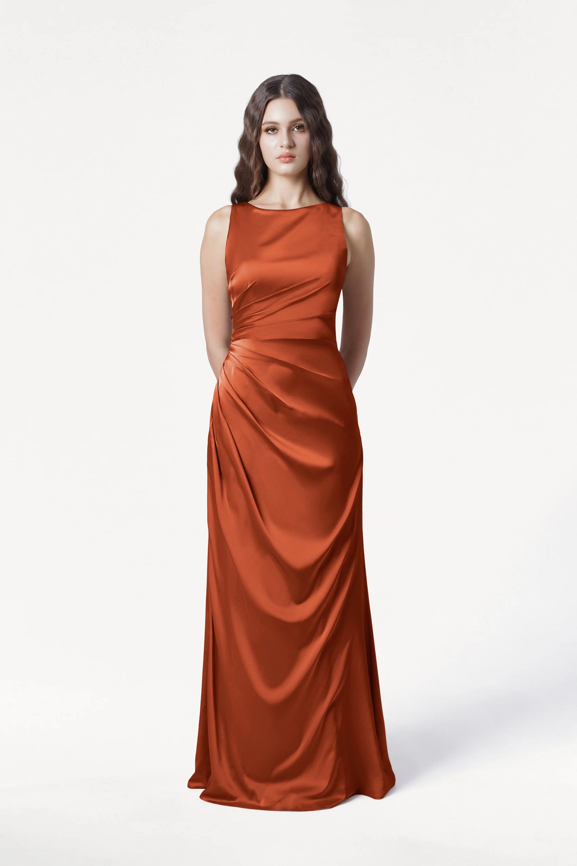 A woman with long, wavy brown hair stands facing forward in the TH&TH Nala Bridesmaid Dress in Terracotta Satin—a sleeveless, floor-length gown—on a plain white background. Perfect for modern bridal parties.