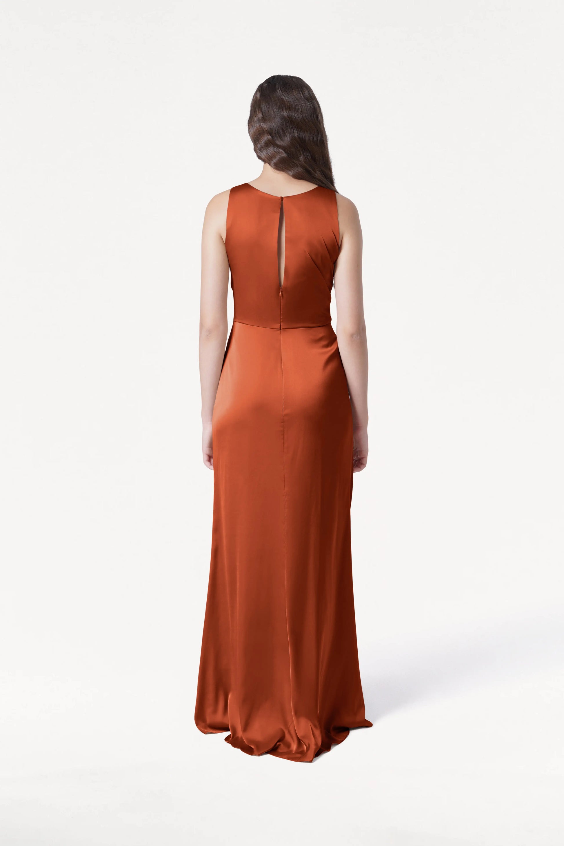 A woman with long brown hair faces away, wearing the TH&TH Nala Bridesmaid Dress in Terracotta Satin—an elegant, sleeveless, floor-length choice for modern bridal parties—shown against a plain white background.