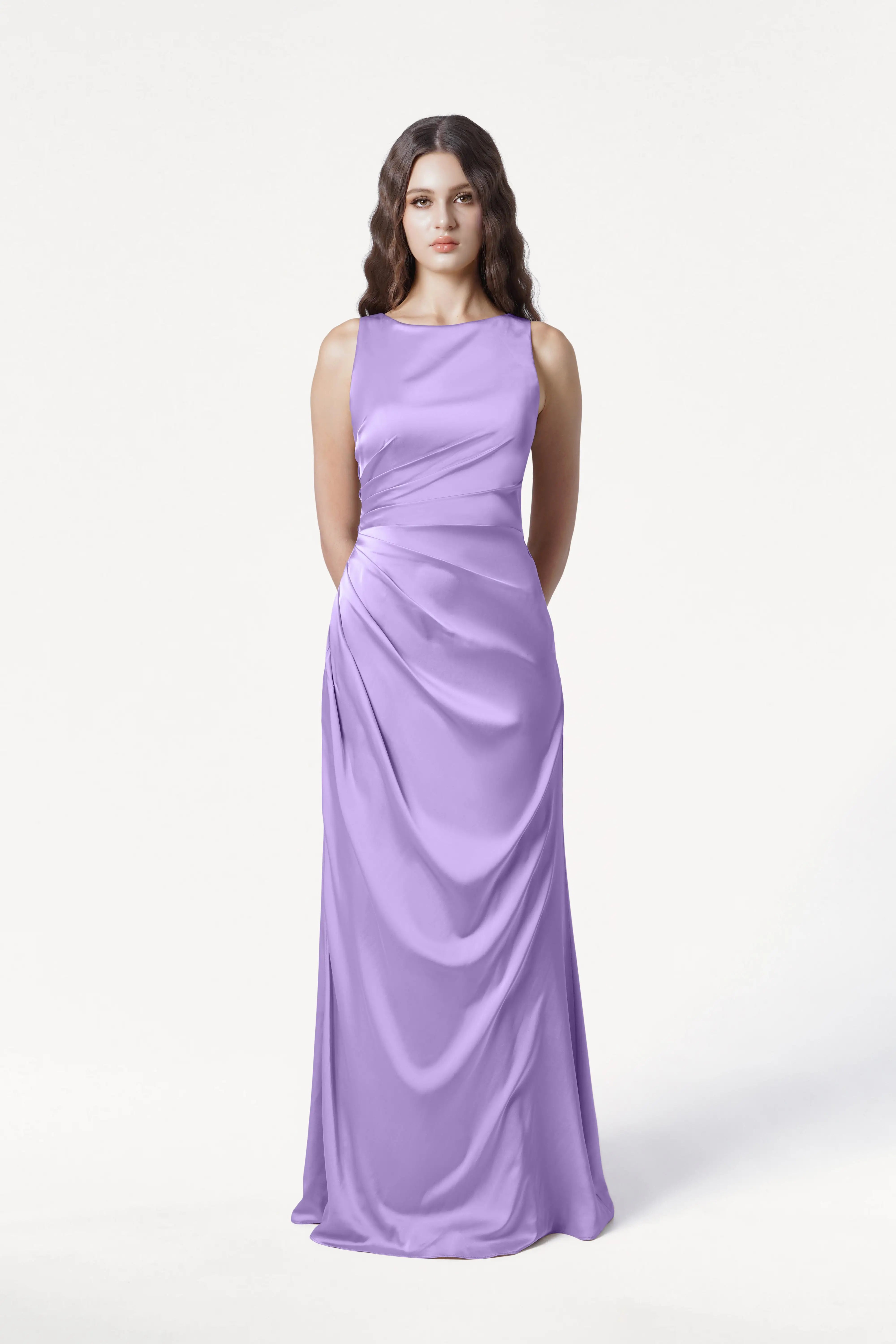 A woman with long wavy brown hair stands against a plain white background, wearing the TH&TH Nala Bridesmaid Dress in Violet Satin, featuring Grecian-inspired pleating and draping. Her hands are held behind her back—a modern, elegant look for bridal parties.