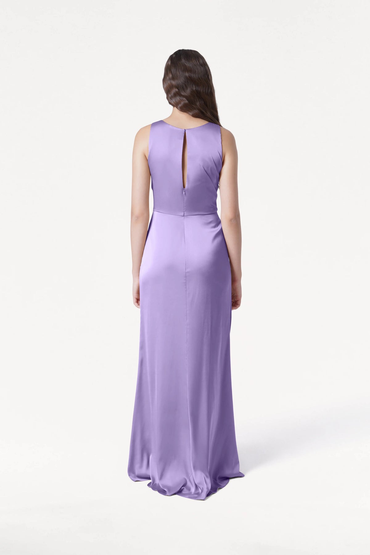 A woman with long brown hair, seen from behind, wears the TH&TH Nala Bridesmaid Dress in violet satin—a floor-length, sleeveless gown with Grecian-style pleating and a small back slit. The background is plain white.