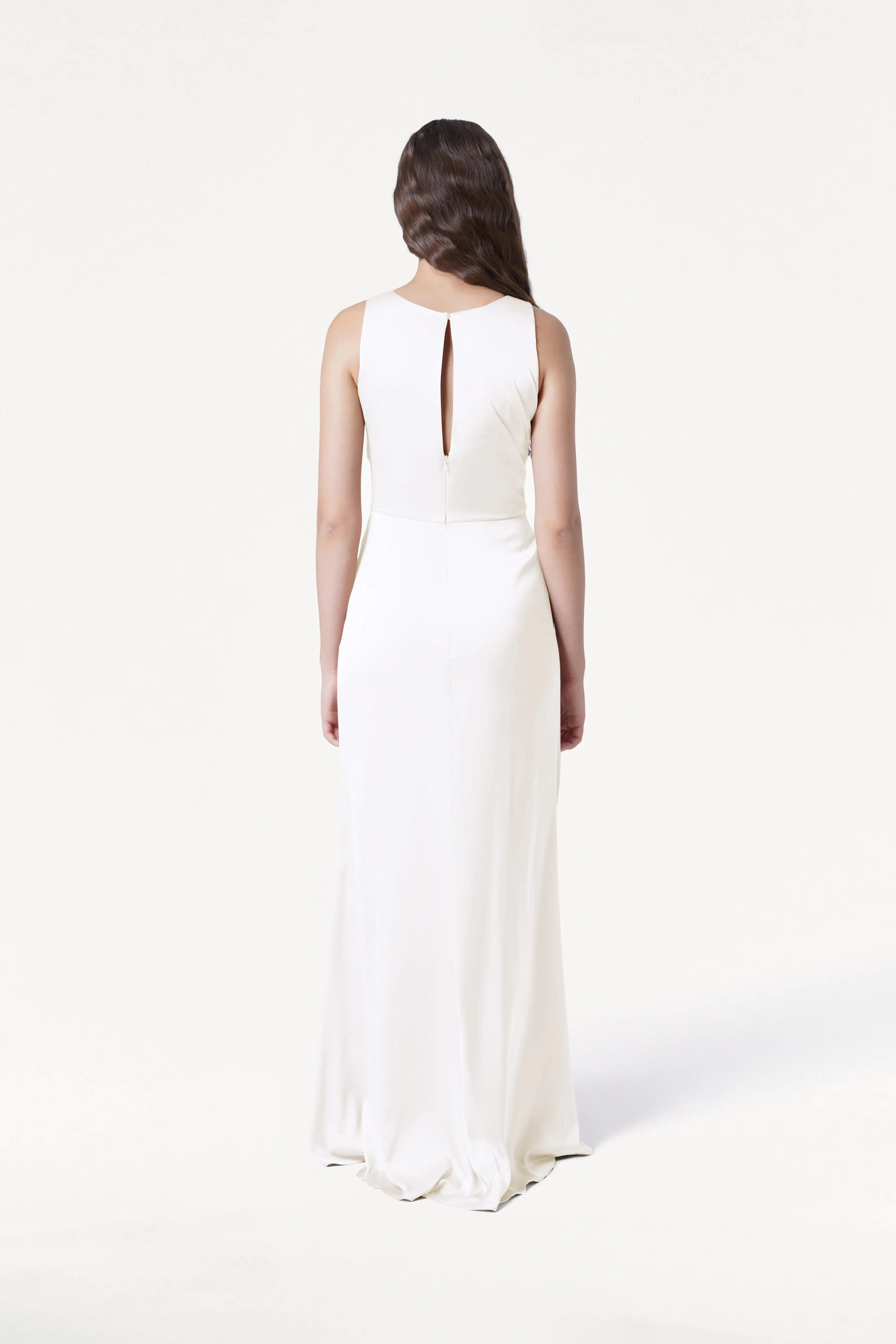 A woman with long, wavy hair stands facing away in the TH&TH Nala Dress in Ivory Satin, featuring Grecian-inspired pleating and a small keyhole at the upper back, set against a plain white background.