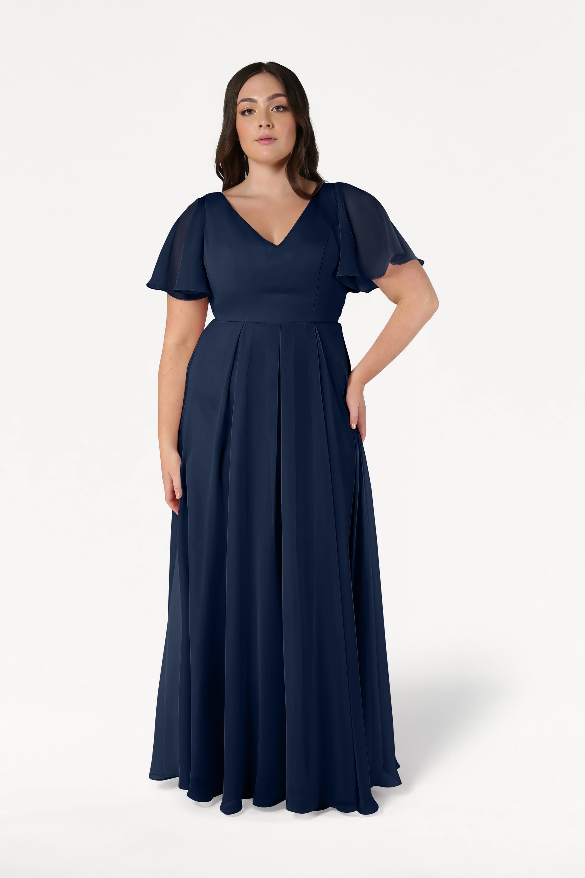 A woman with loose waves and a neutral expression models the TH&TH Phoebe Bridesmaid Dress in Navy Chiffon, featuring a floor-length pleated skirt, V-neckline, and short flutter sleeves against a white background—ideal for bridesmaids.