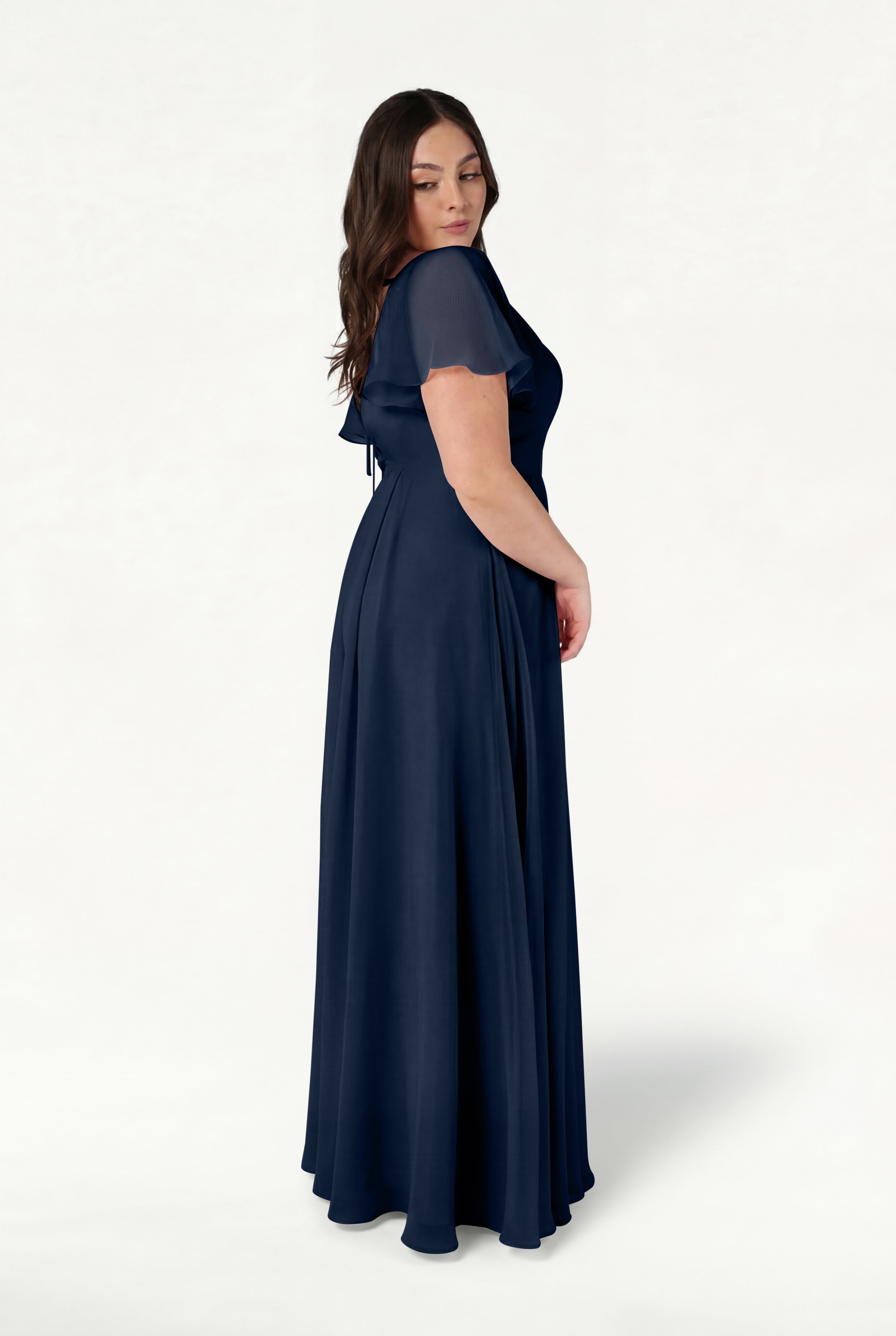 A woman with long brown hair stands in profile against a white background wearing the TH&TH Phoebe Bridesmaid Dress in navy chiffon, featuring short sleeves and a flowing silhouette.