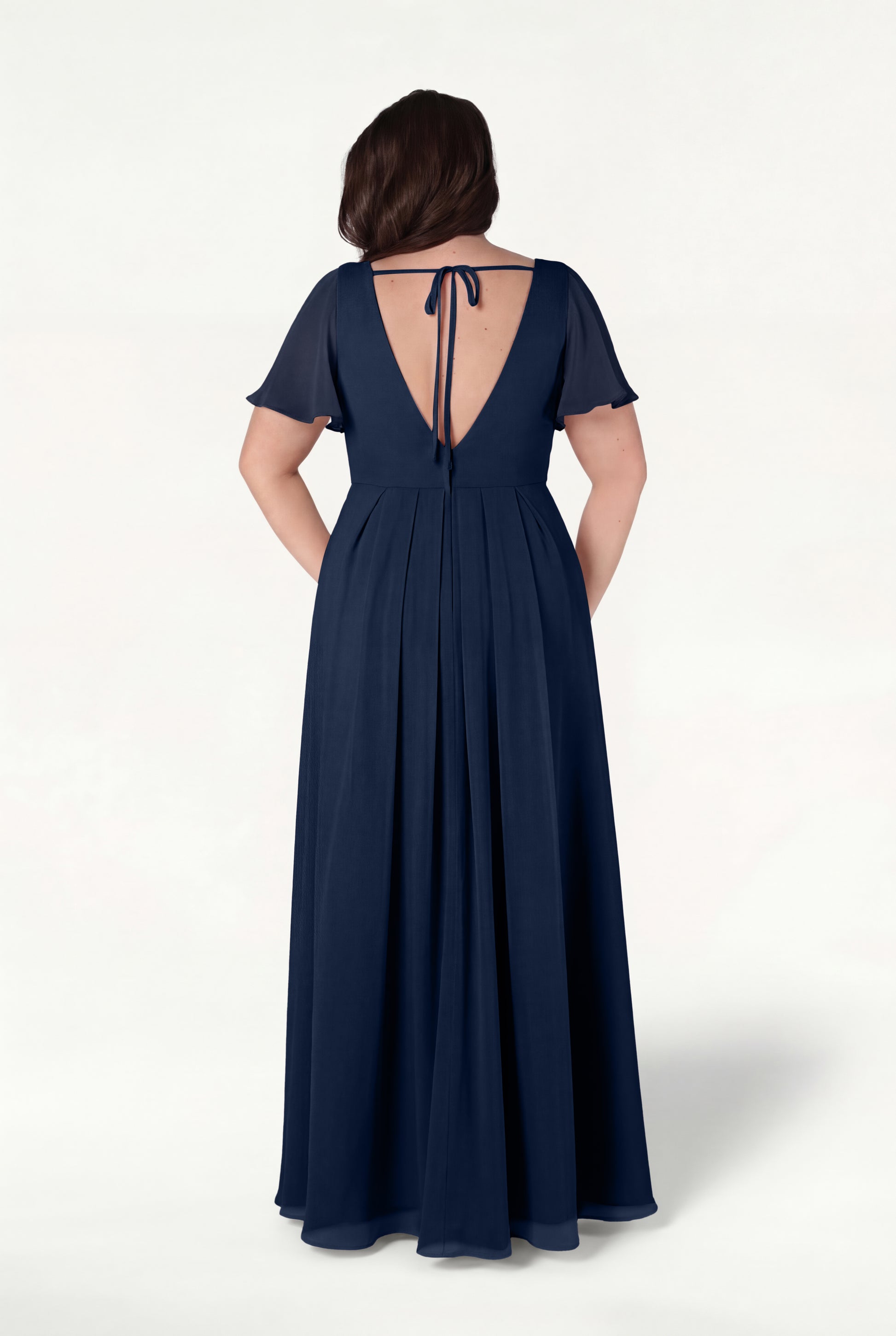 A woman is seen from the back in the TH&TH Phoebe Bridesmaid Dress in Navy Chiffon—a floor-length, short-sleeve gown featuring a V-shaped open back with a tie and a flowing, bump-friendly skirt.