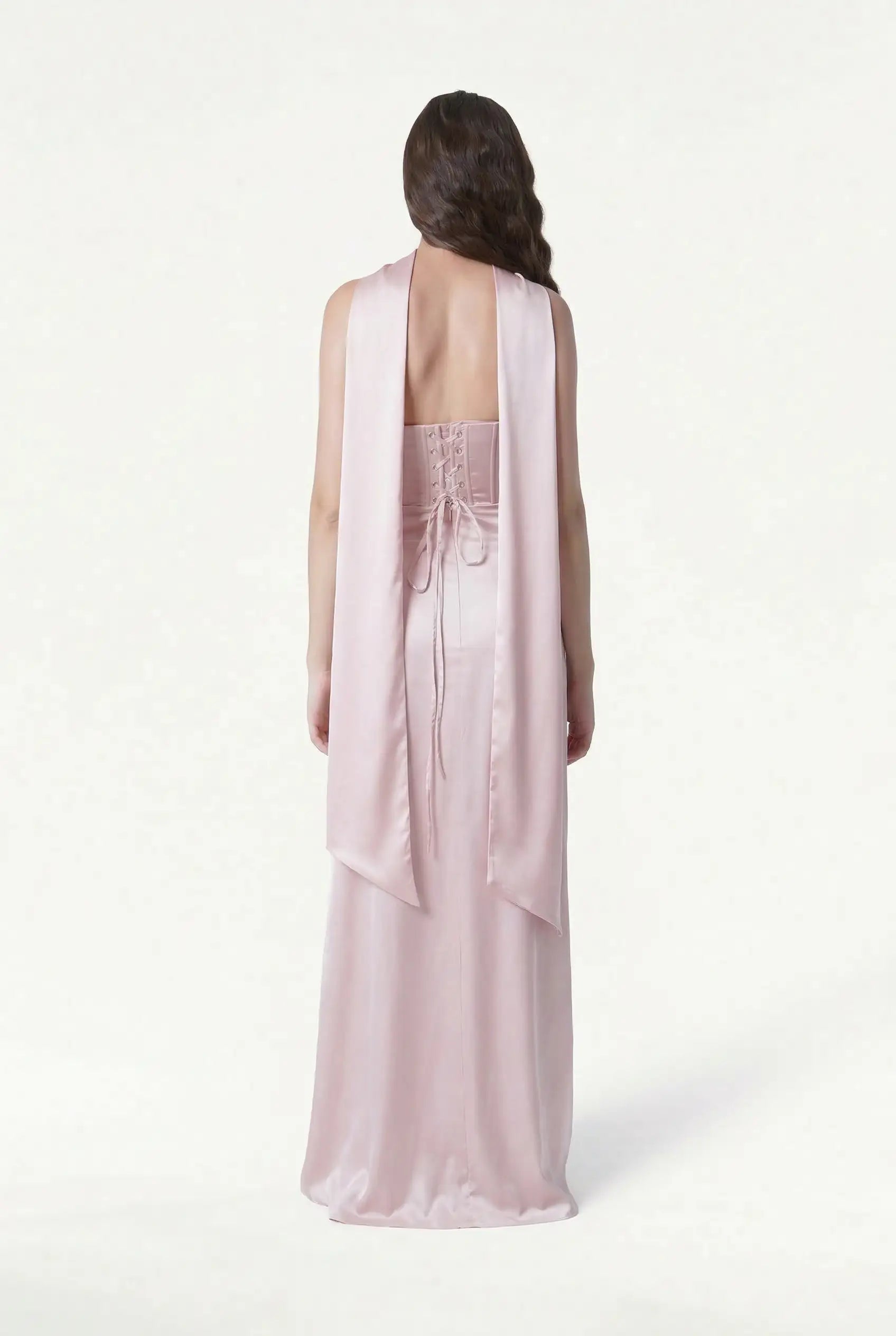 A woman with loose brown waves wears TH&TH’s Neck Scarf & Sash in light pink satin, styled as a sash with lace-up details. She faces away against a plain white background.