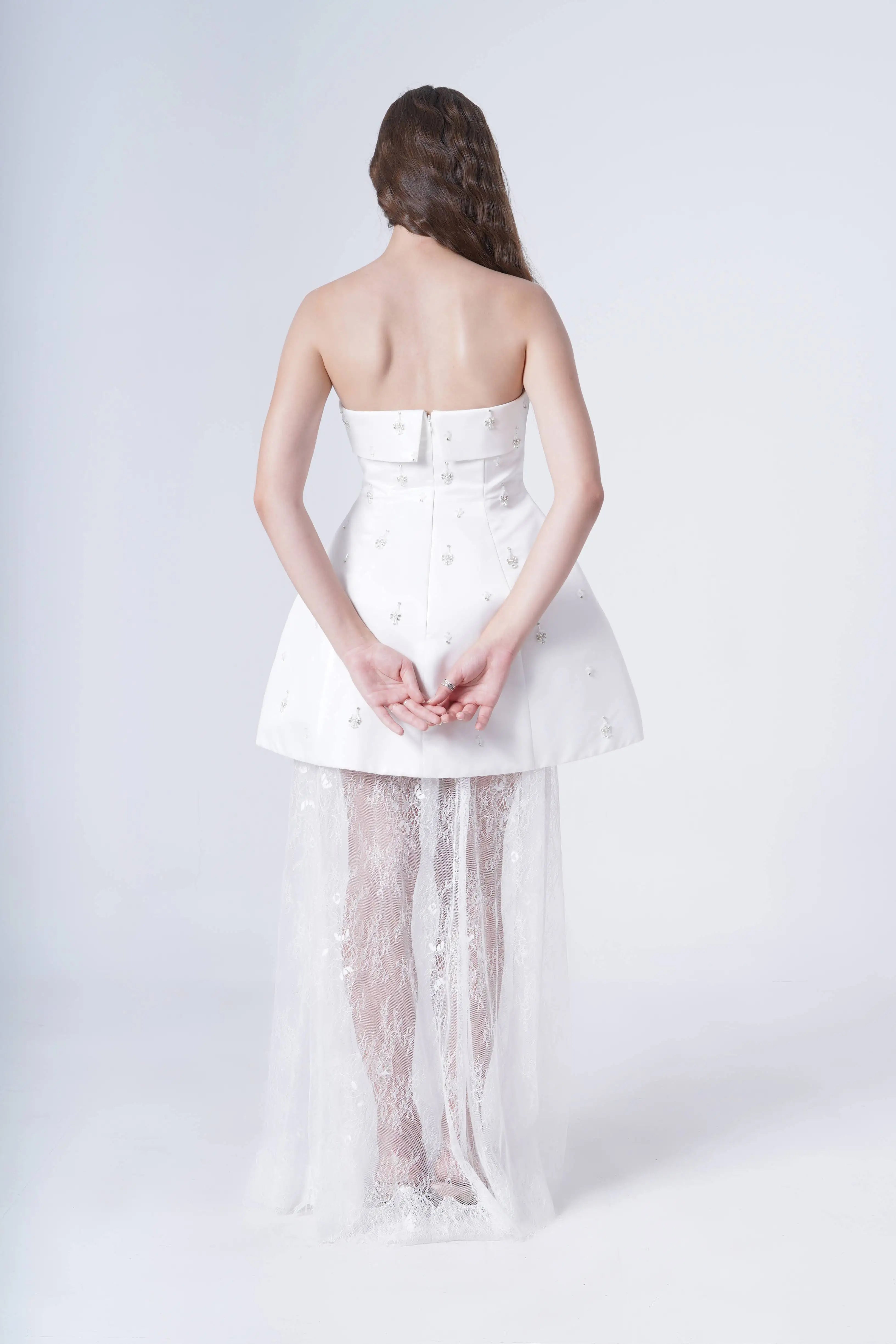 A woman with long brown hair stands facing away, hands clasped behind her back, wearing the TH&TH Nicholl Bridal Skirt in Ivory Chantilly Lace—strapless, short, with a sheer lace overlay flowing to the floor.