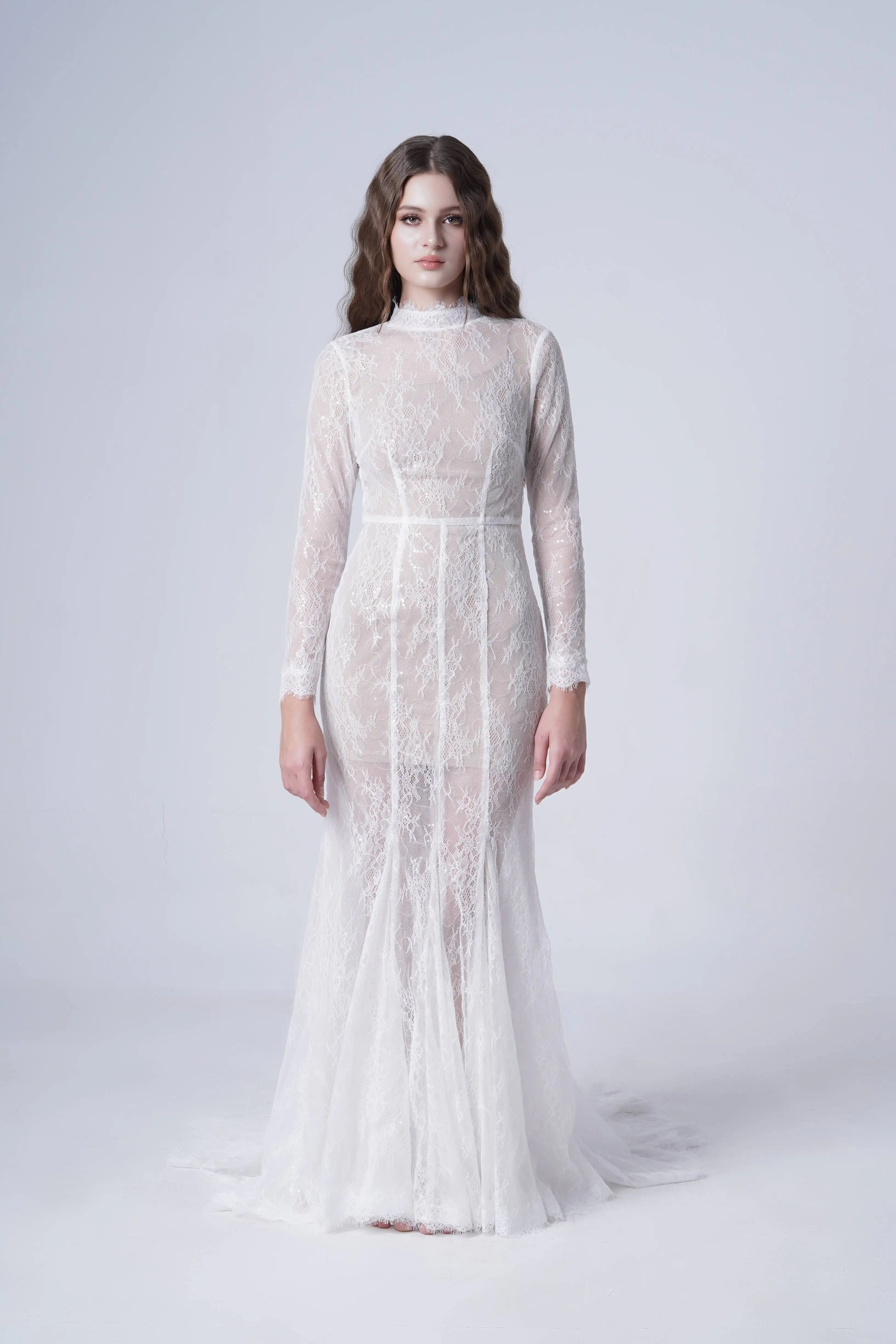 A woman faces forward in the Nova Bridal Dress in Ivory Chantilly Lace by TH&TH—a floor-length, sheer white lace gown with long sleeves and a high neckline. Her hair is in loose waves against a plain light gray background.