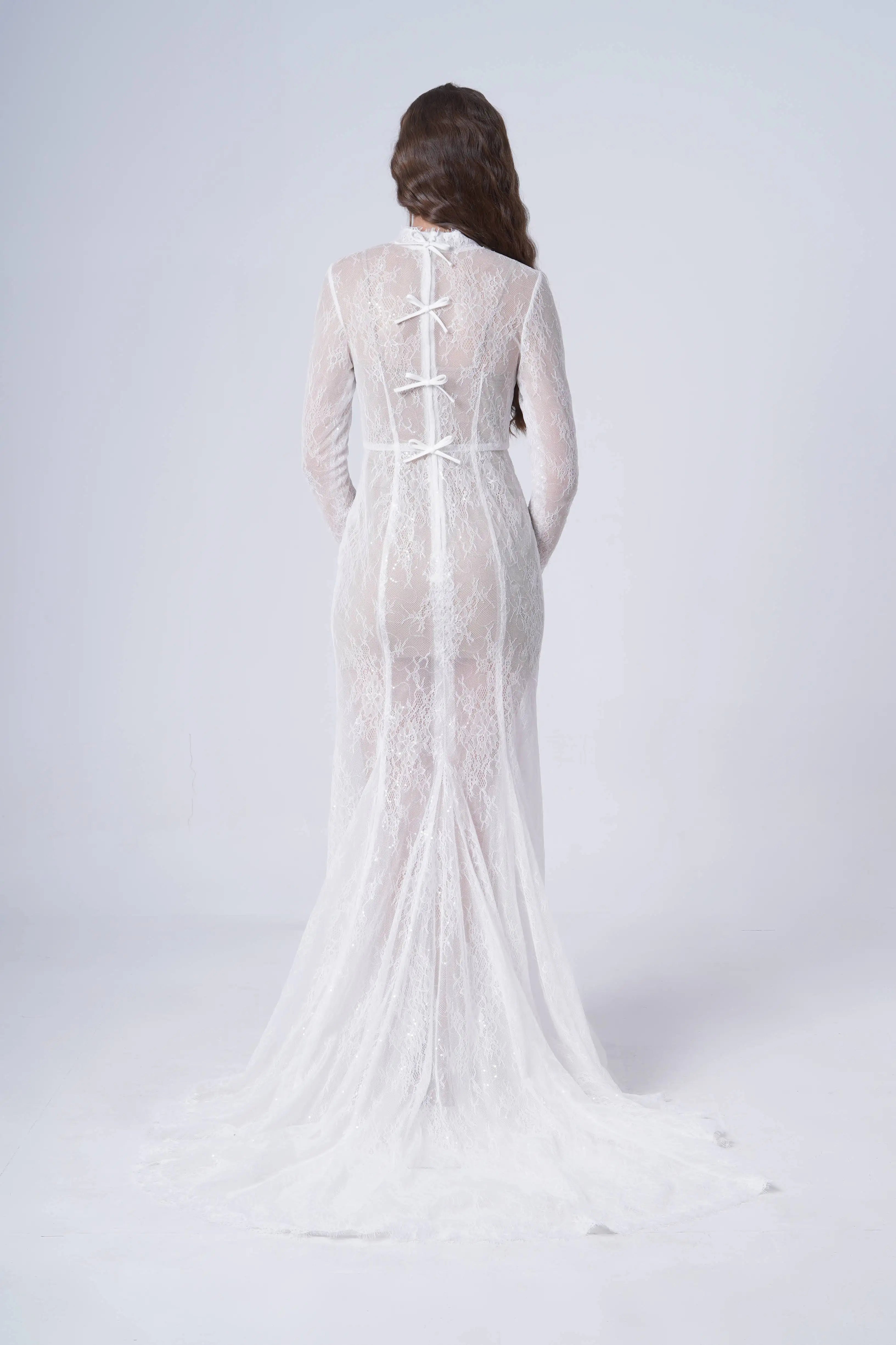 A woman with long brown hair stands with her back to the camera, wearing the TH&TH Nova Bridal Dress in Ivory Chantilly Lace, featuring ribbon ties down the back, against a plain white background.