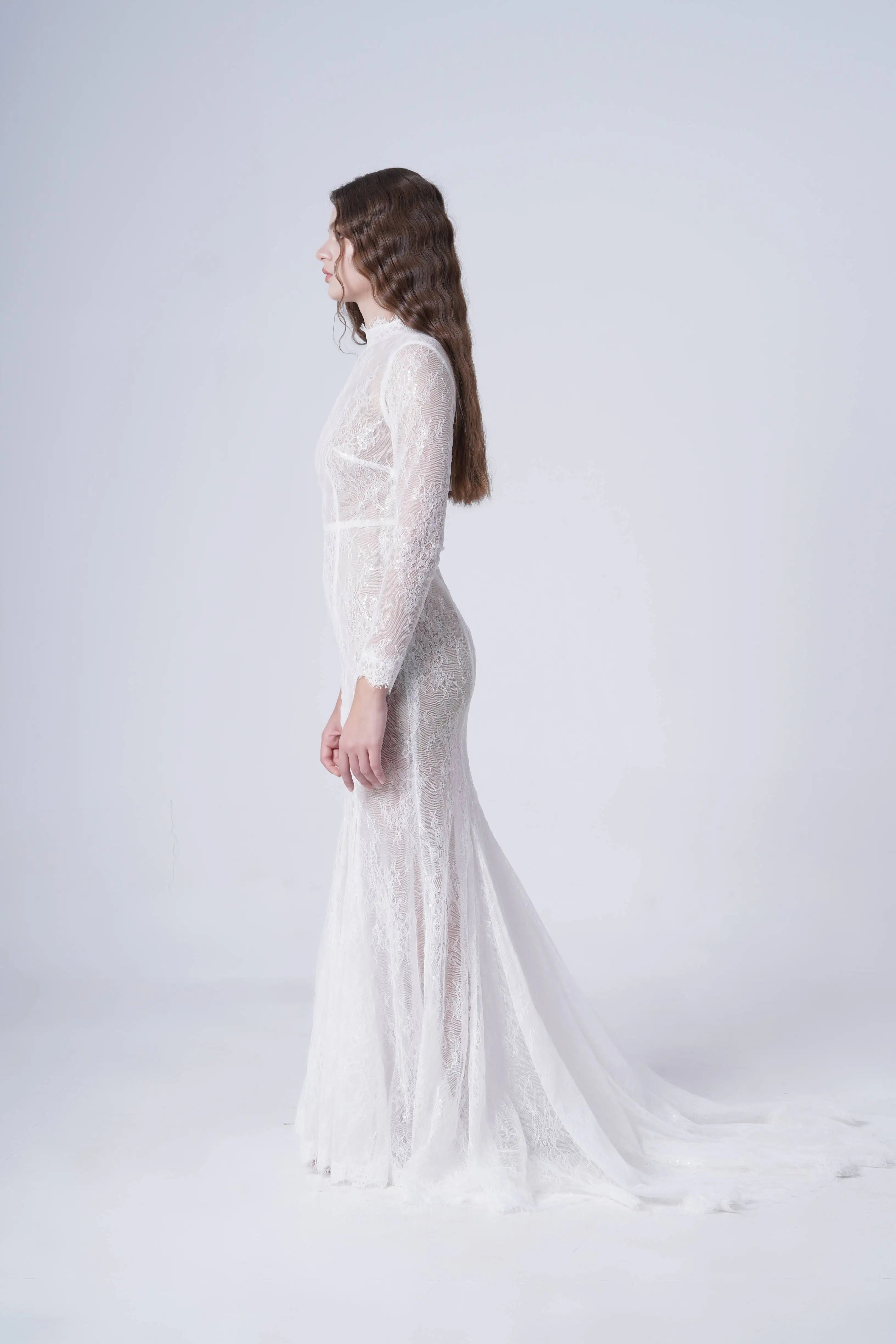 A woman with long wavy hair stands in profile wearing the TH&TH Nova Bridal Dress in ivory Chantilly lace, a sheer floor-length gown with long sleeves and a train, set against a plain light background.