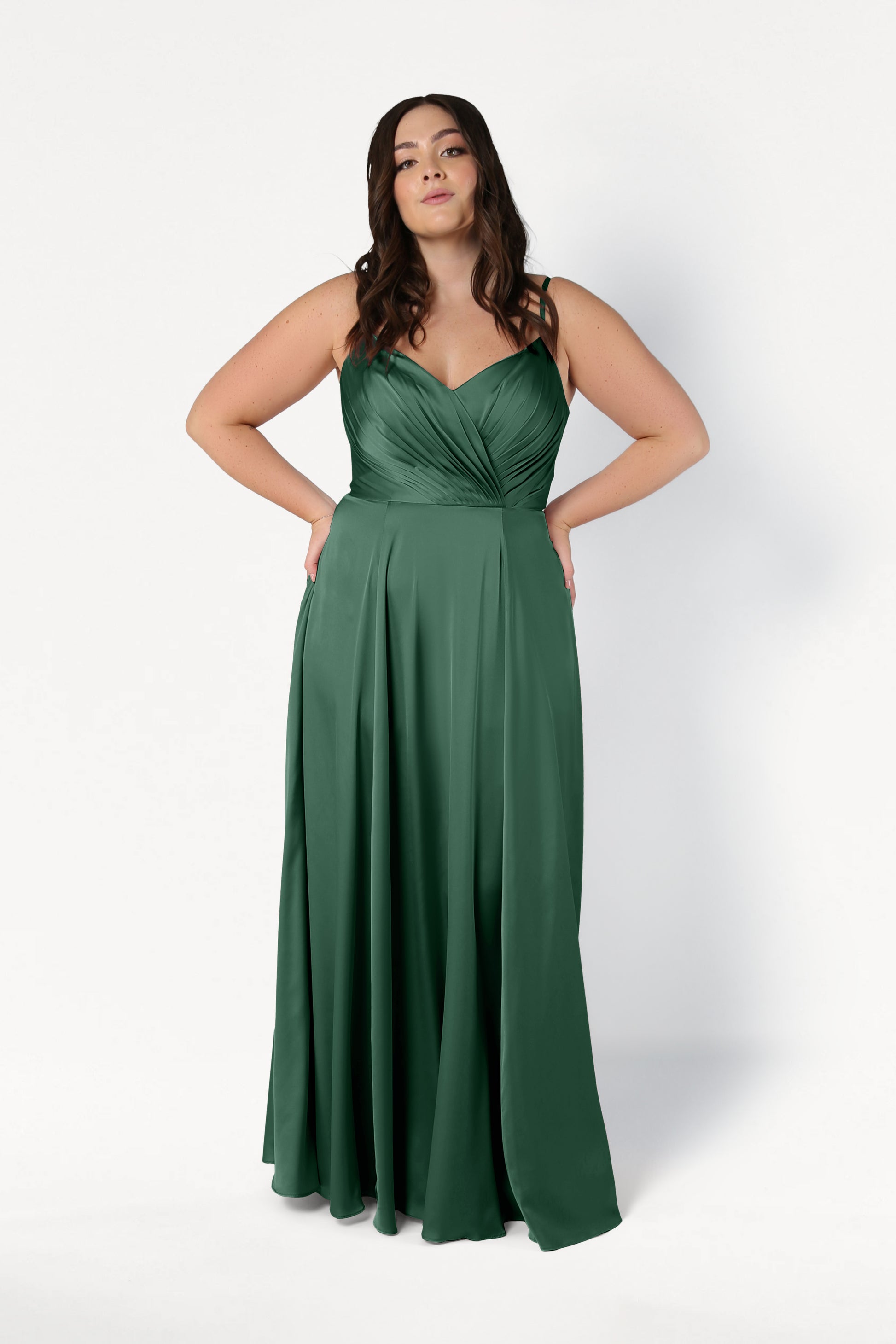 A woman with long brown hair wears the TH&TH Isla Bridesmaid Dress in Olive Grove Satin, a sleeveless, floor-length gown with a fitted pleated bodice and flowing skirt, standing against a plain white background.