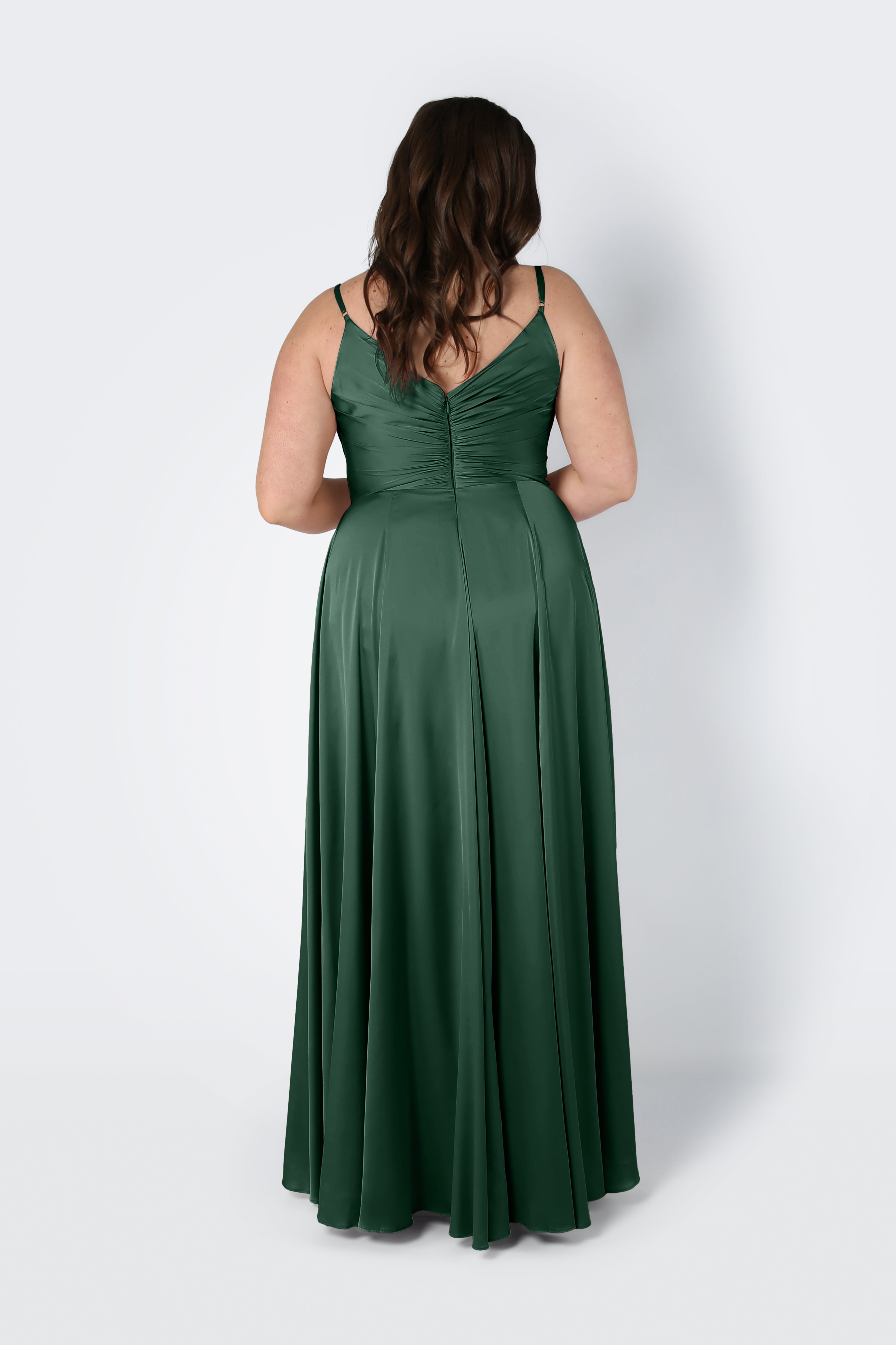 A woman with wavy brown hair stands with her back to the camera against a plain light background, wearing the TH&TH Isla Bridesmaid Dress in Olive Grove Satin, featuring thin straps and a pleated bodice.