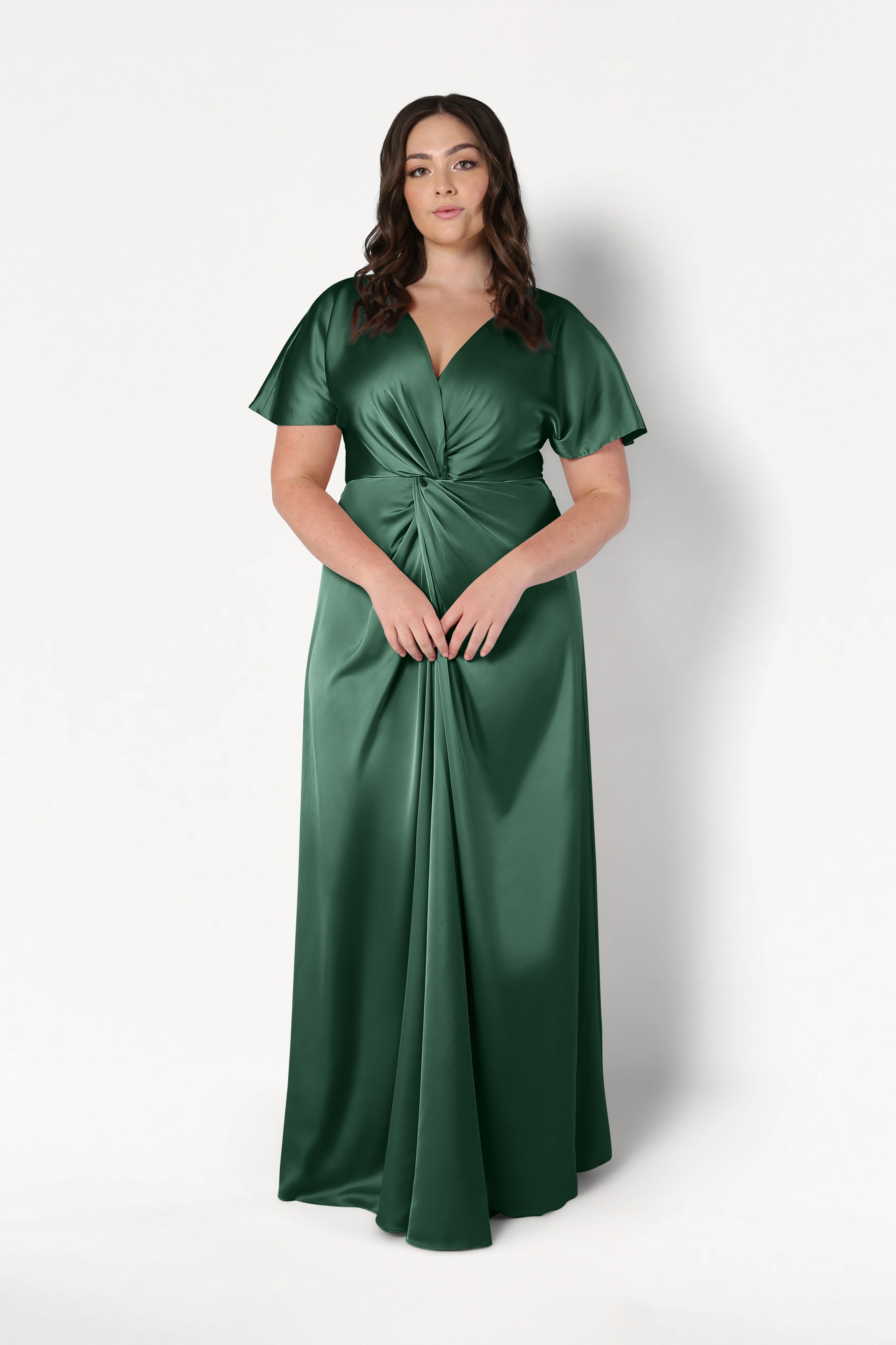 A woman with long, wavy brown hair and a neutral expression wears the TH&TH Camilla Bridesmaid Dress in Olive Grove Satin—a floor-length gown with short flutter sleeves, a V-neckline, and a gathered knot waist—against a white background.