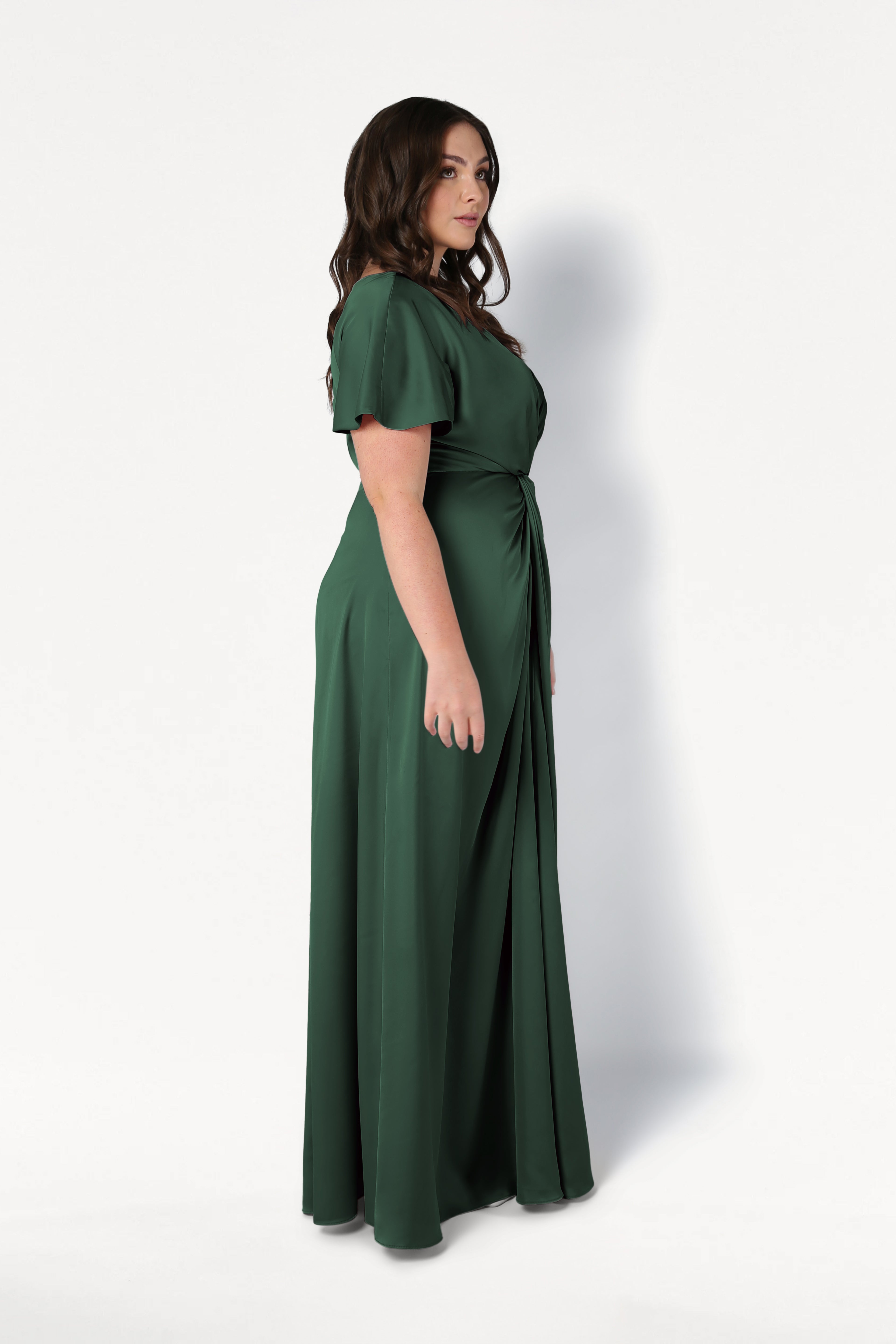 A woman with wavy brown hair stands in profile, modeling the TH&TH Camilla Bridesmaid Dress in Olive Grove Satin—a modern, floor-length gown with short sleeves and a gathered waist—against a plain white background.