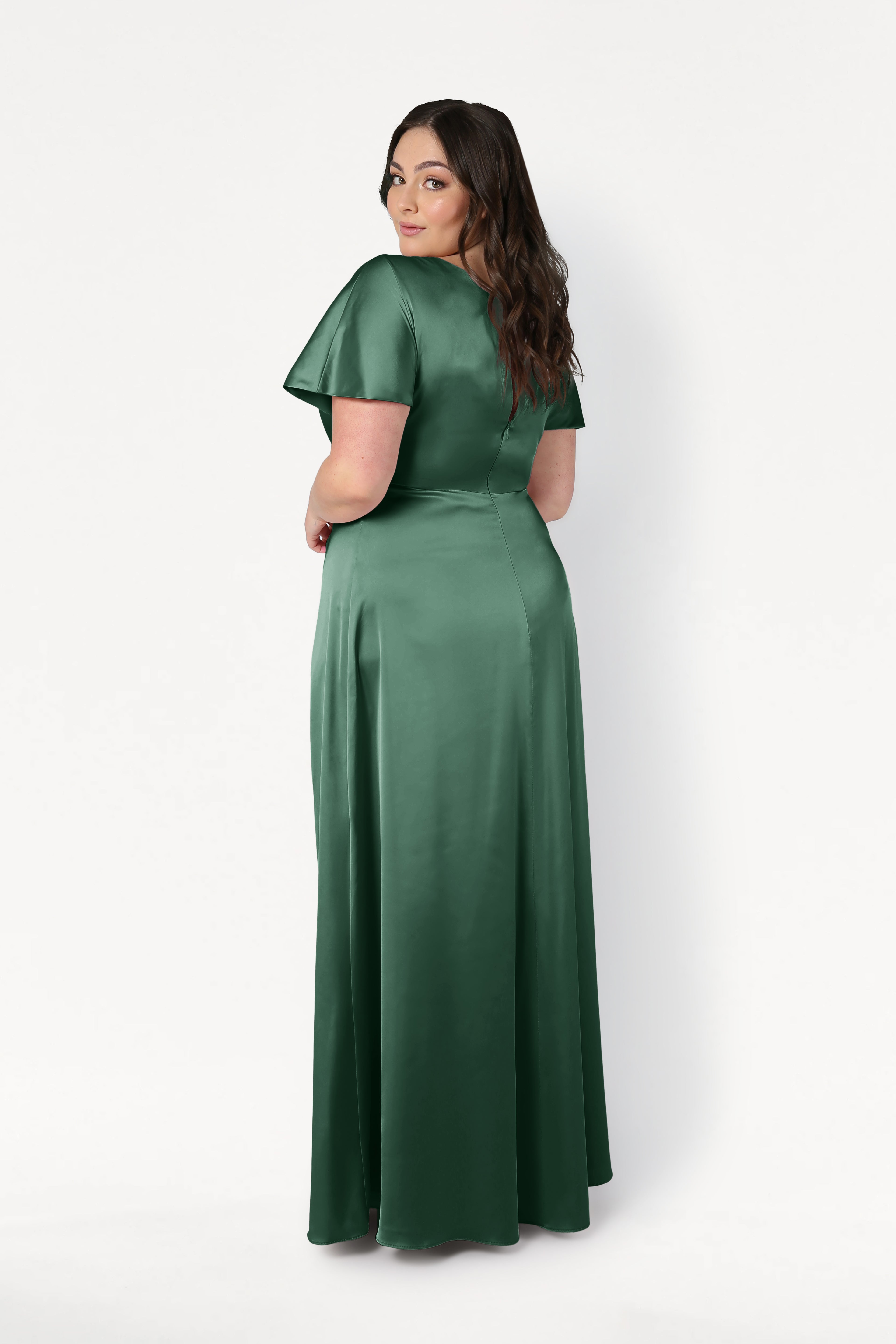 A woman with long brown hair wears the TH&TH Camilla Bridesmaid Dress in Olive Grove Satin, a floor-length, short-sleeve gown. She stands against a plain white background, facing away and looking back over her shoulder.