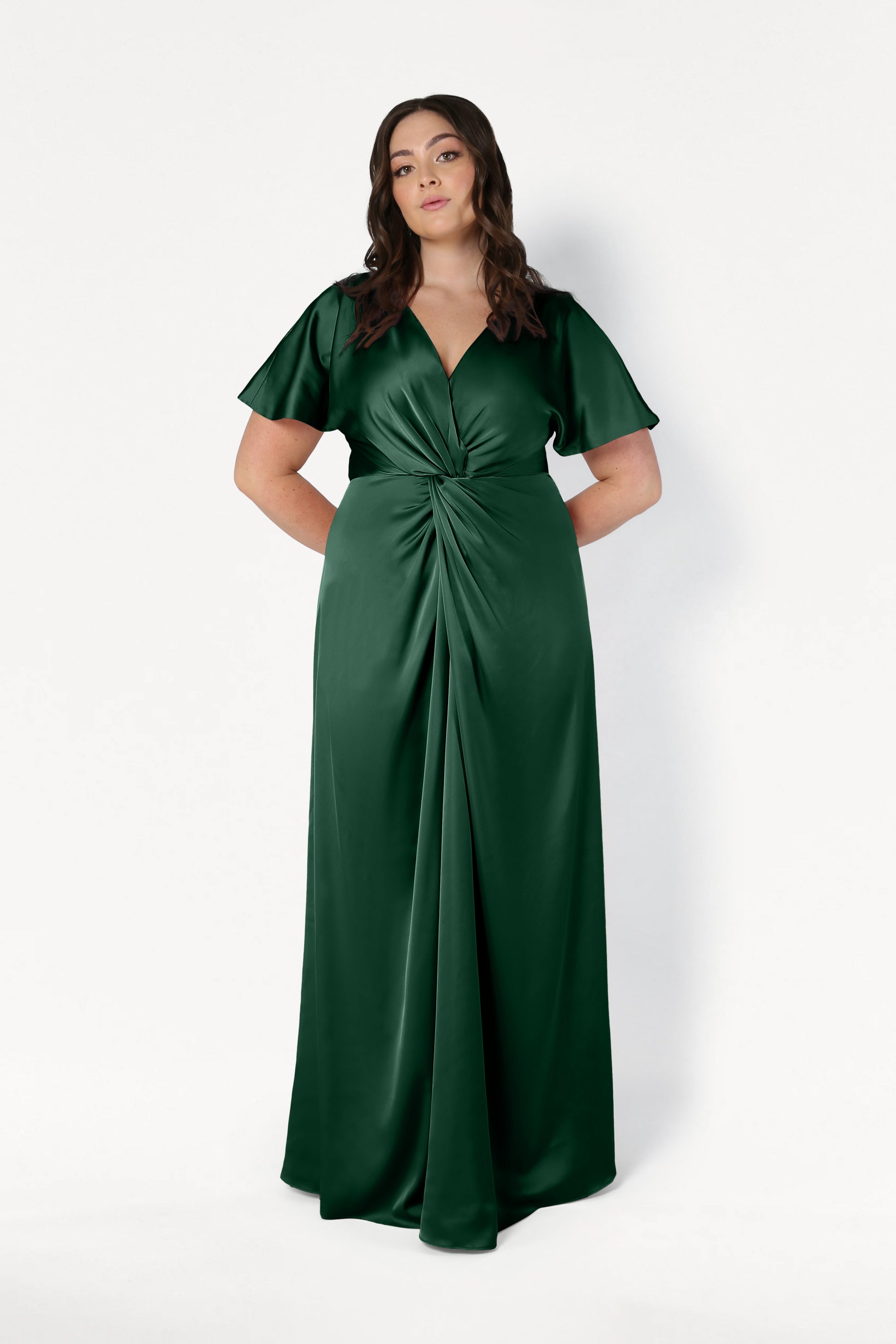A woman with long brown hair wears the Camilla Bridesmaid Dress in Olive Grove Satin by TH&TH, featuring short flutter sleeves and a front knot detail, standing against a white background with her hands behind her back.