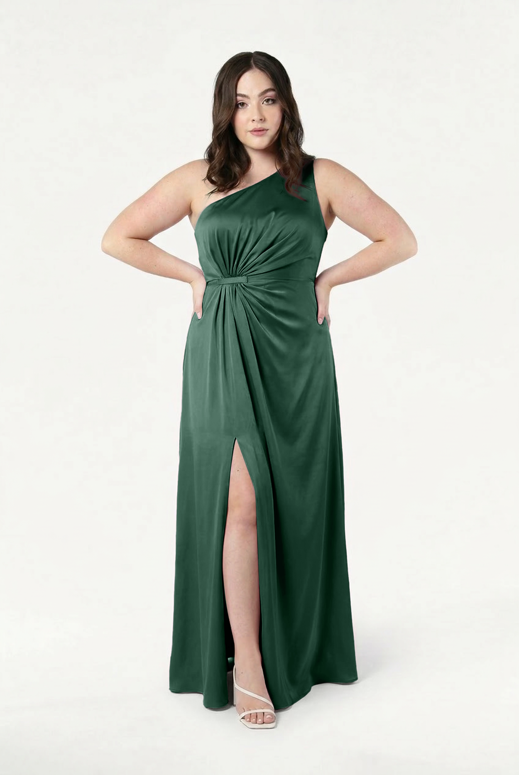 A woman wears the TH&TH Greta Bridesmaid Dress in Olive Grove Satin—a sleeveless, floor-length gown with Grecian draping and a thigh-high slit—paired with white strappy heels, standing against a plain white background.