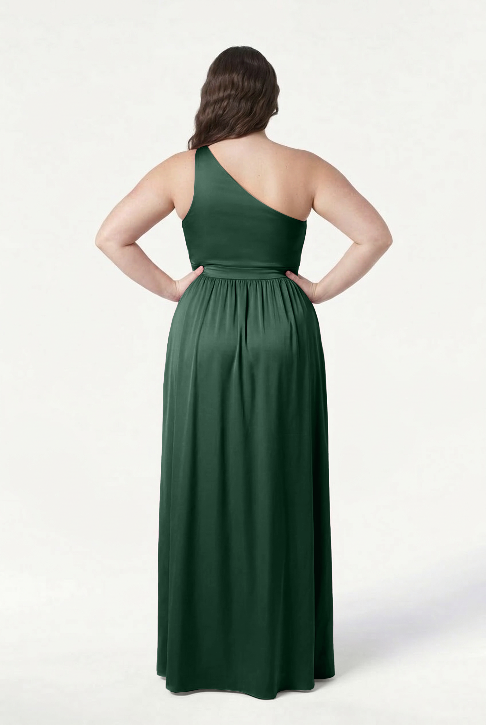 A woman stands with her back to the camera, wearing the TH&TH Greta Bridesmaid Dress in Olive Grove Satin. Her hands rest on her hips and her dark hair cascades in waves past her shoulders against a plain white background.