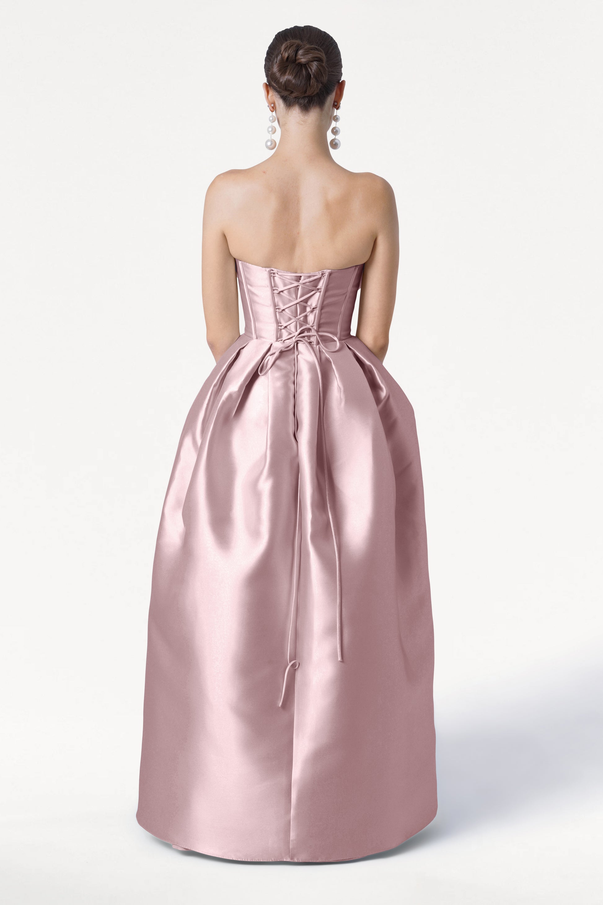 A woman with a neat bun and long, dangling earrings stands with her back to the camera, wearing the TH&TH Ophelia Bridesmaid Dress in blush pink satin twill, featuring a strapless design and lace-up corset bodice.